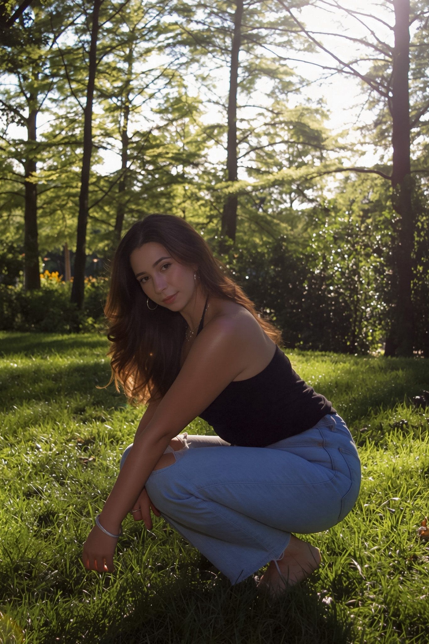 Young woman in black tank top and jeans crouching on grass in sunlit park with trees in background.