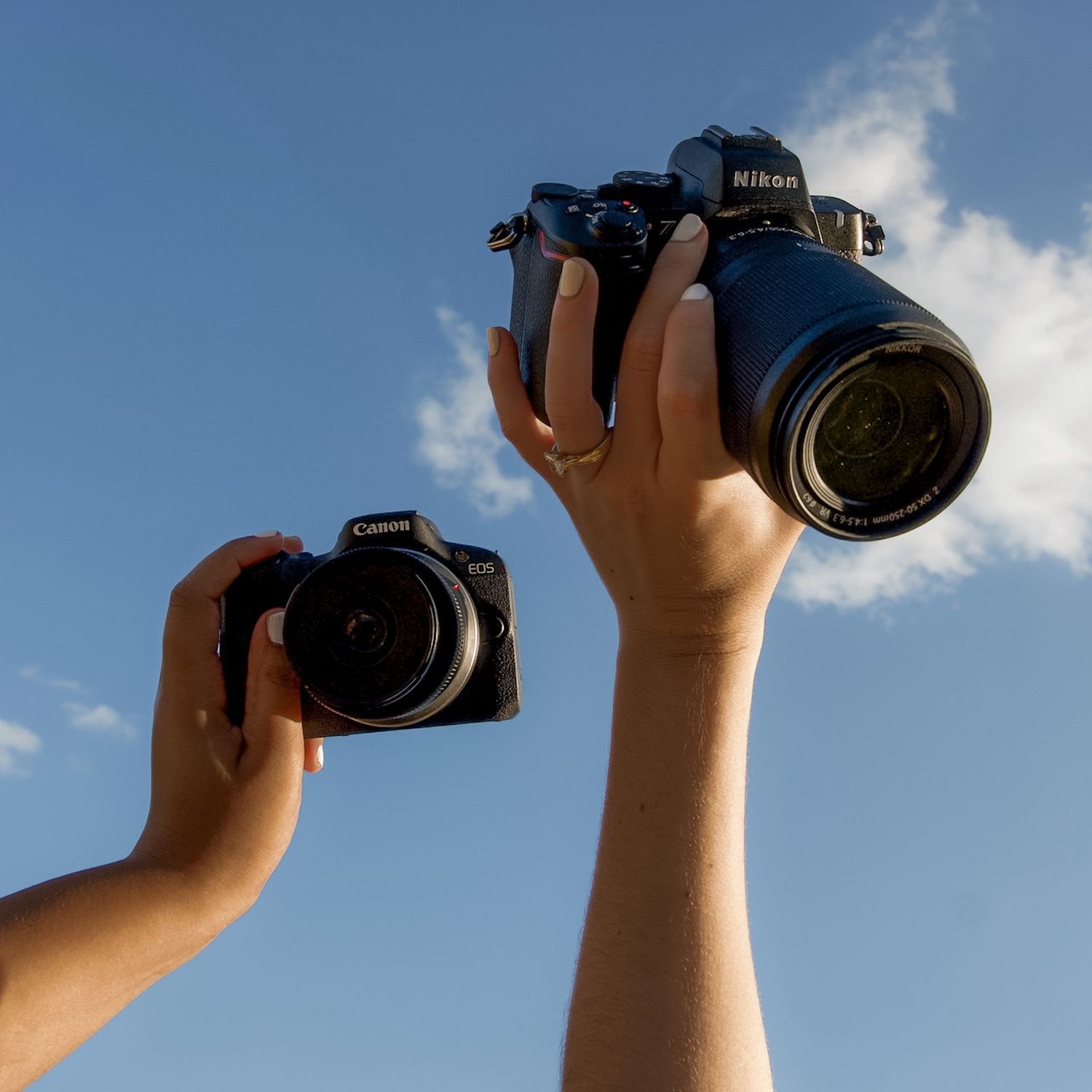 Two hands raised holding a Canon and a Nikon DSLR camera against a bright blue sky.