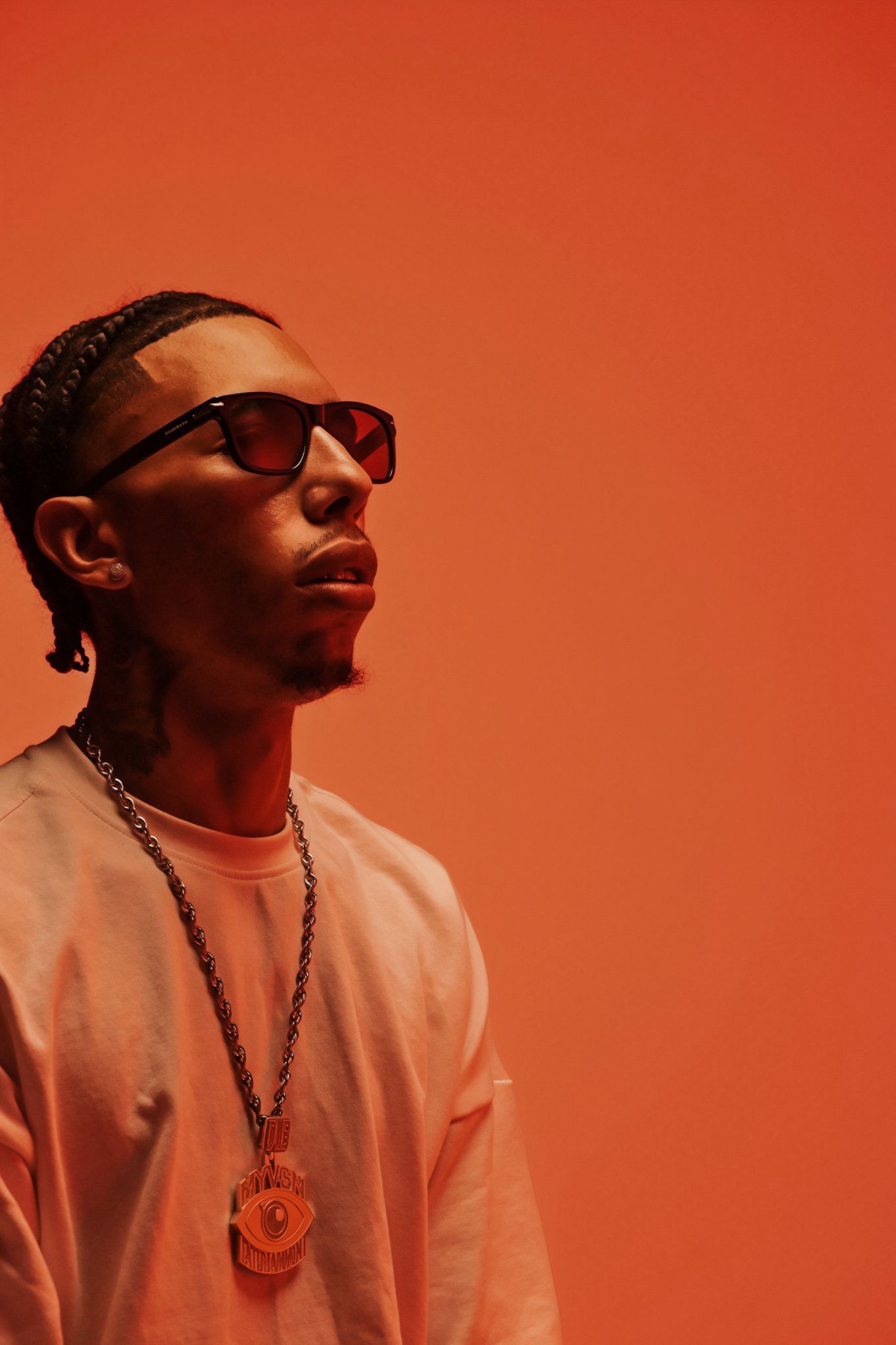 Young Black man wearing sunglasses and a chain pendant posing against a vibrant orange backdrop.