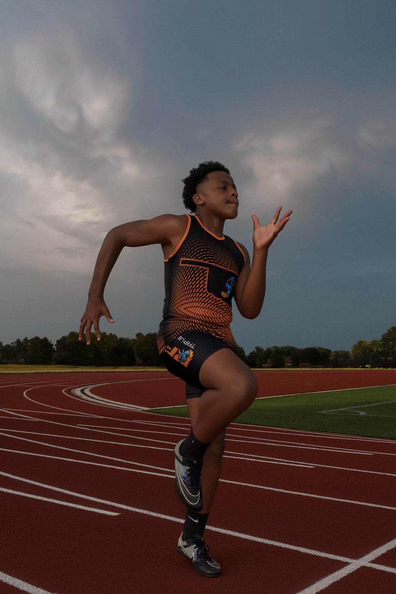 Young male sprinter in orange track uniform running on red track under dramatic cloudy sky.