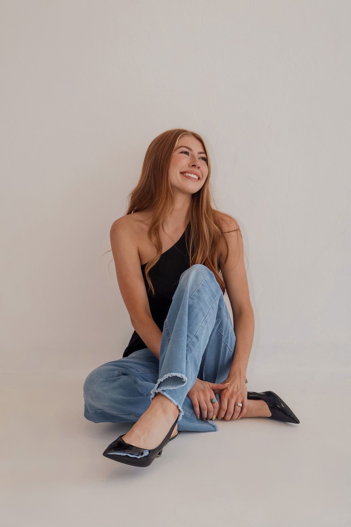 Smiling redhead woman in black top and blue jeans sitting on floor against white background.