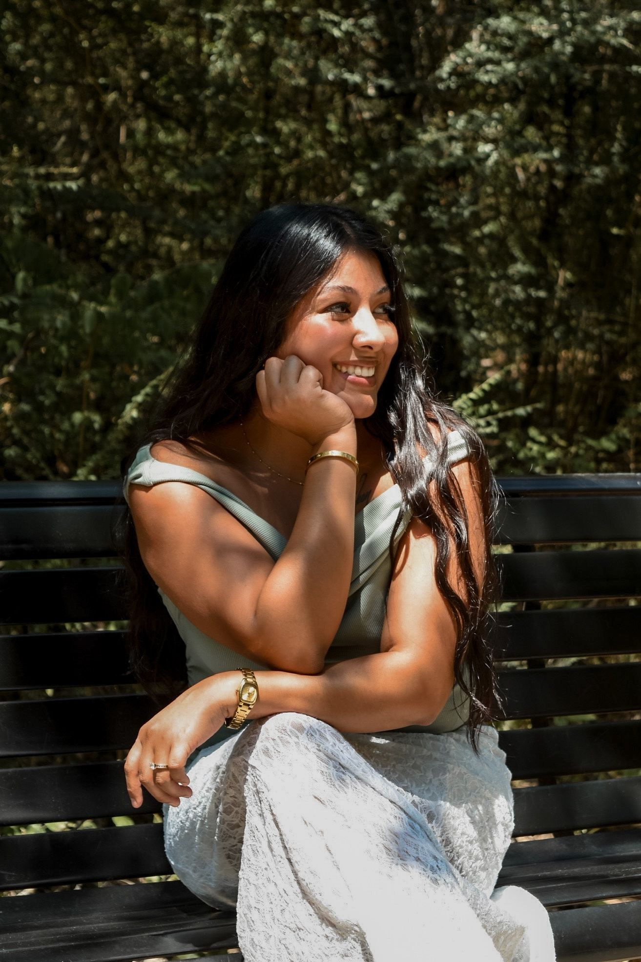 Young woman with long dark hair smiling while sitting on bench in sunny outdoor park setting.