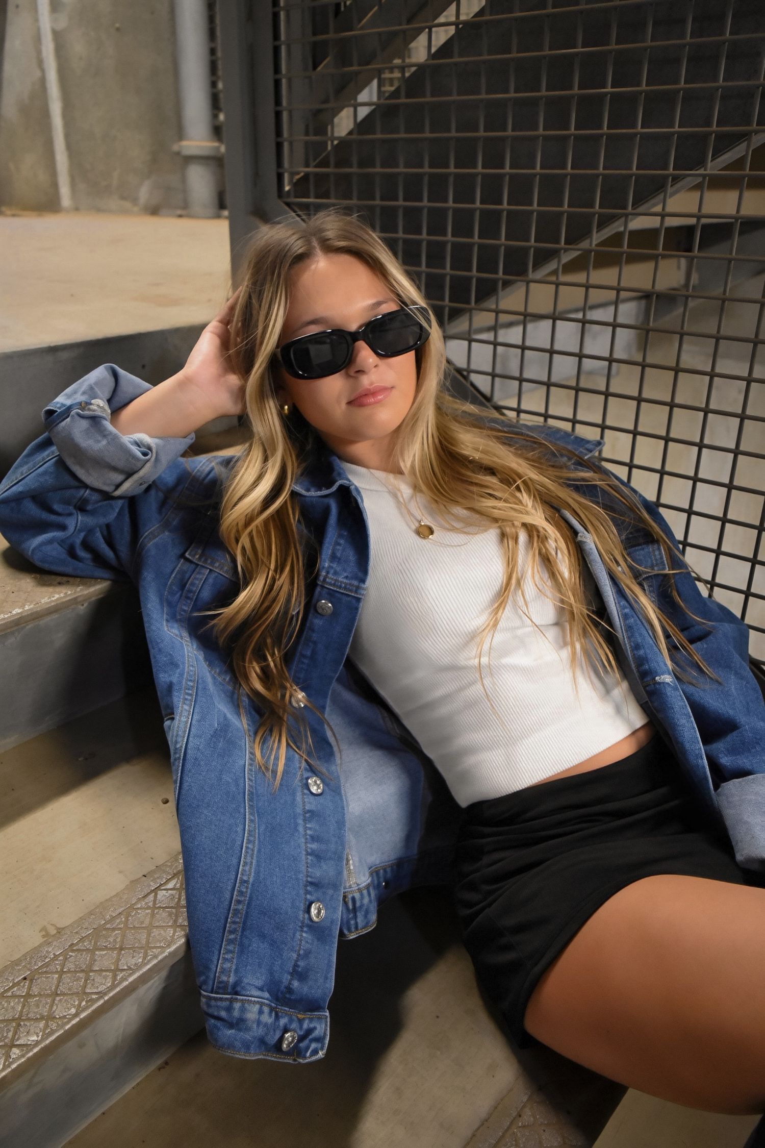 Blonde woman in denim jacket, white crop top and black shorts sitting on steps wearing sunglasses.