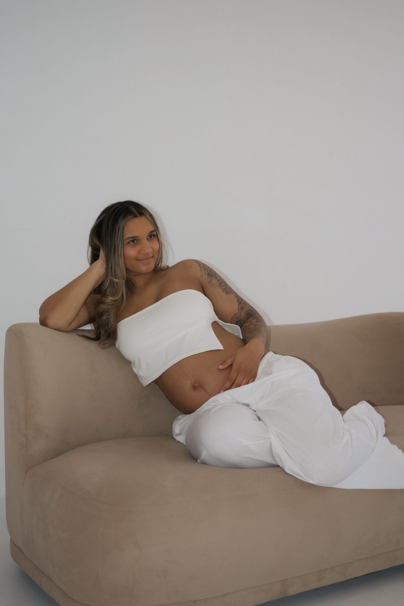 Pregnant woman in white crop top and pants relaxing on beige sofa against white background.
