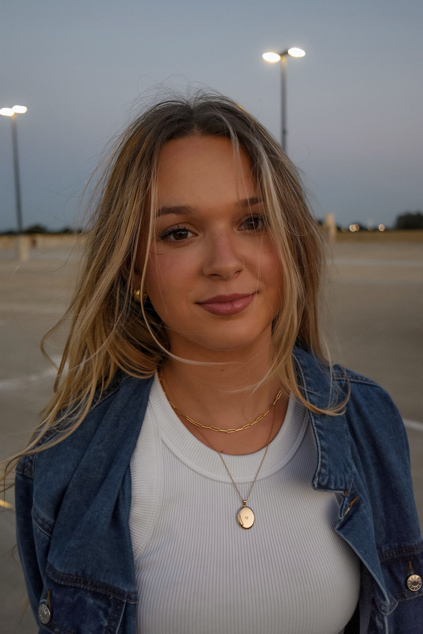 Young blonde woman in denim jacket and gold necklaces posing in parking lot at dusk.