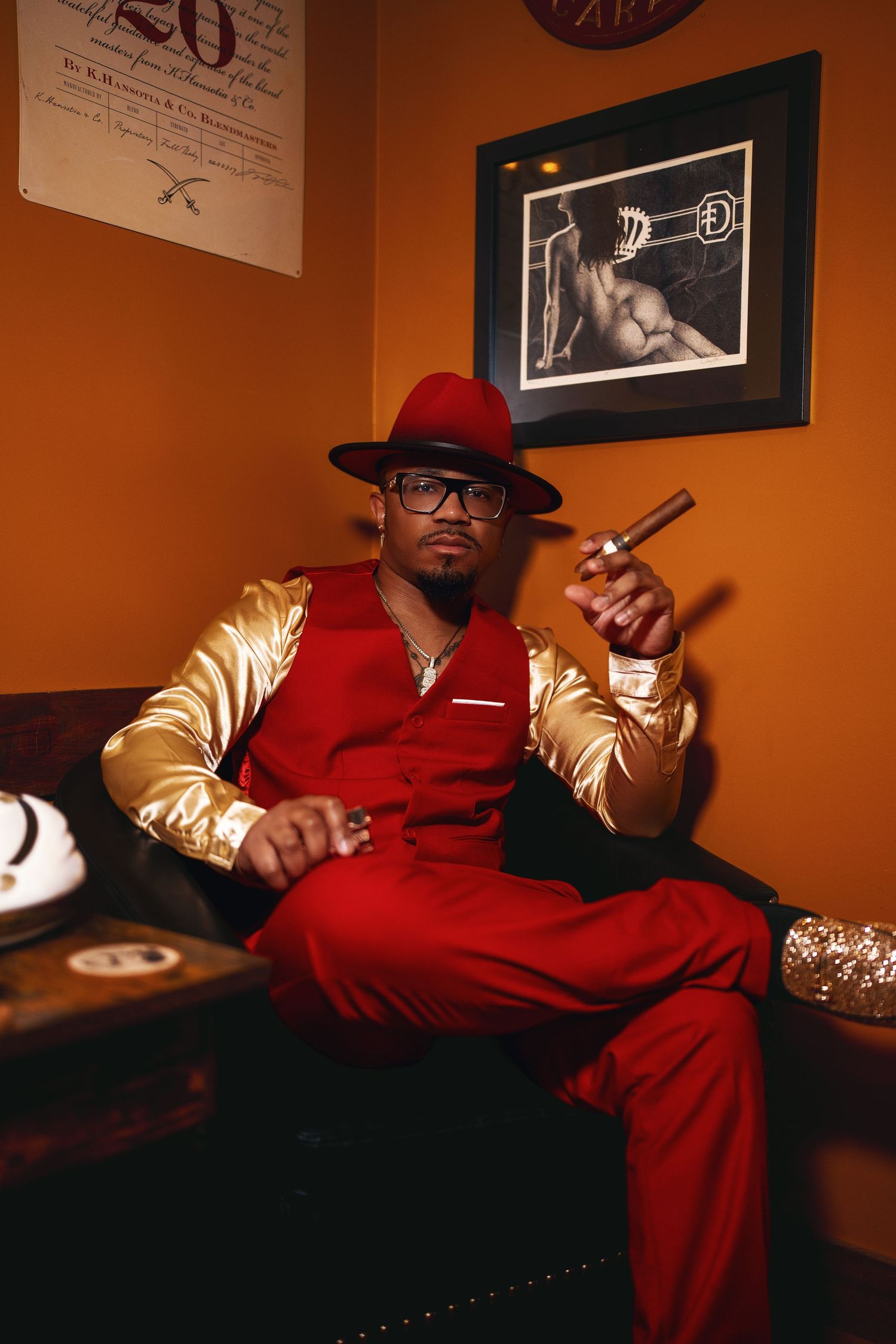 Man in red and gold suit holding a cigar, seated in a warmly lit lounge.