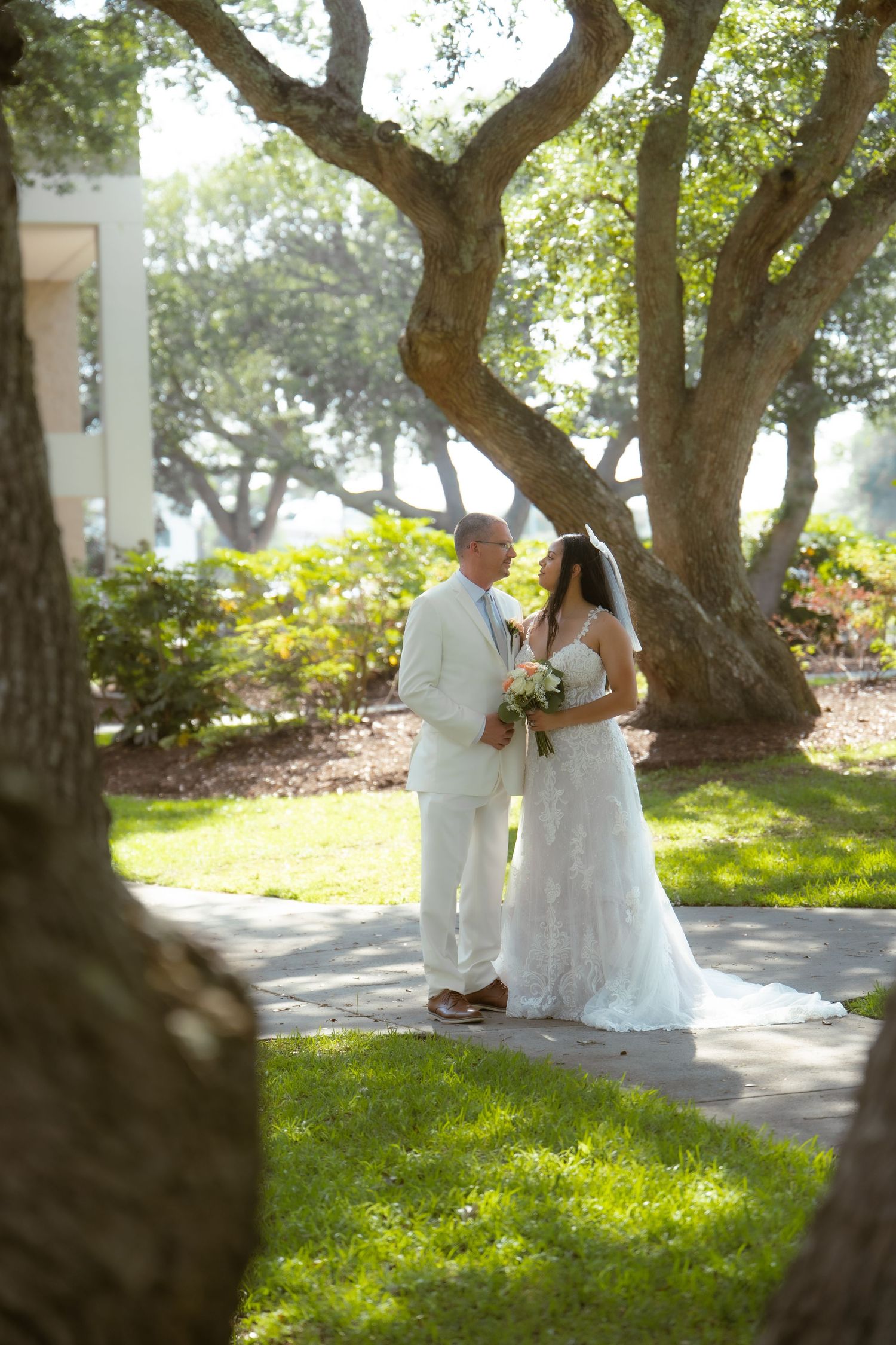 Bride and groom standing together on a garden path beneath large oak trees.