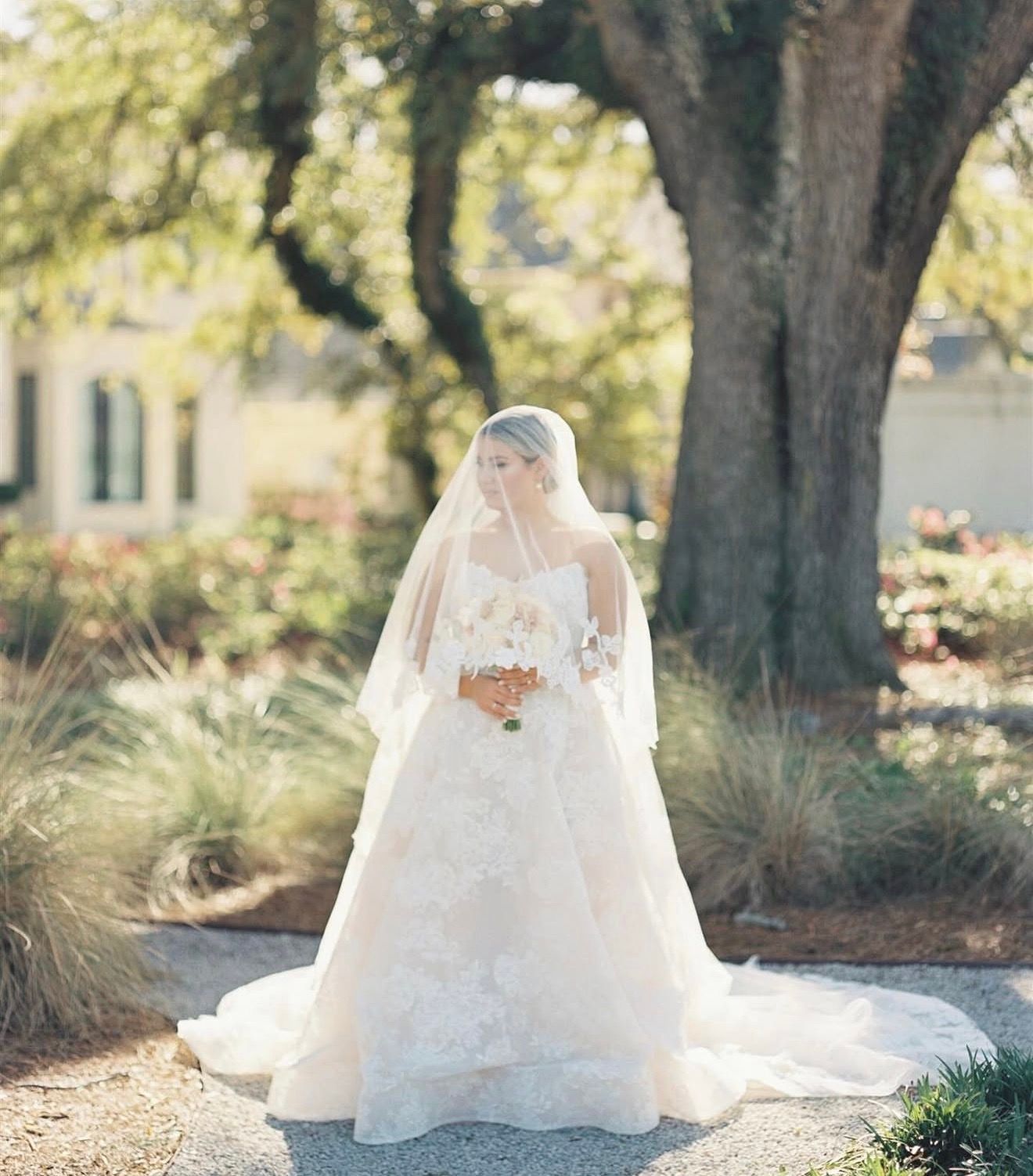 Bride in a layered white gown and veil holding a bouquet under oak trees.