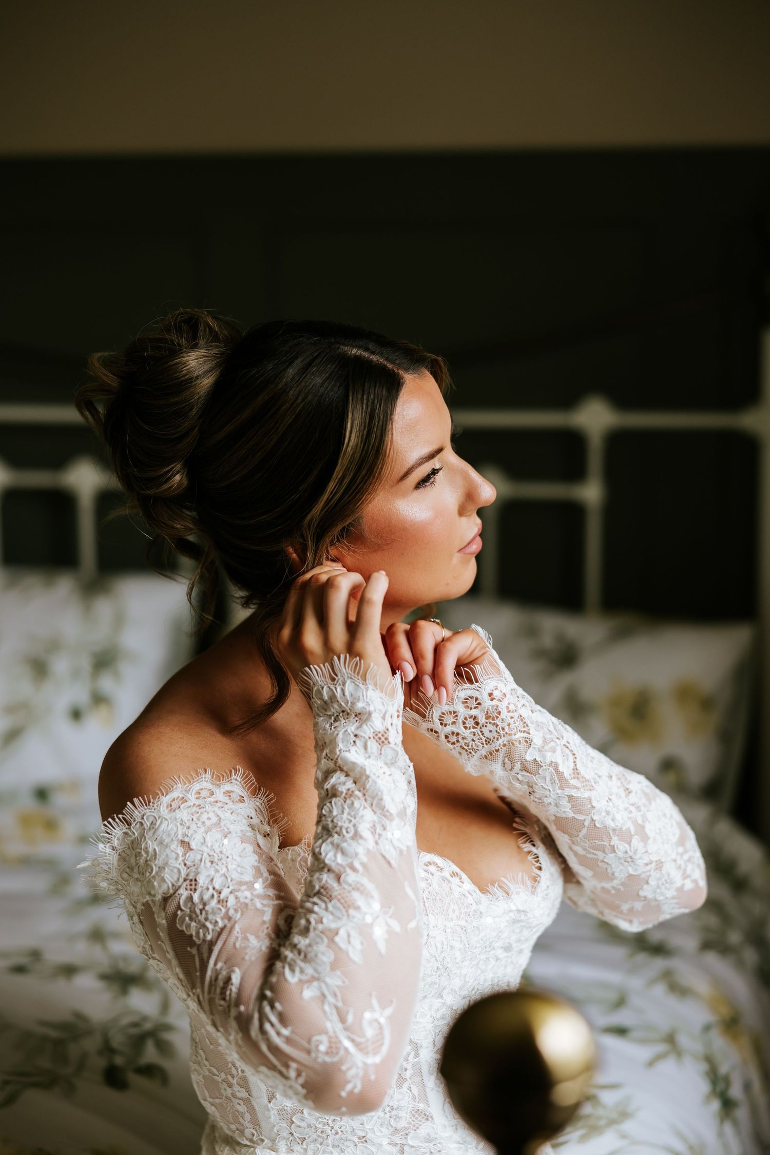 Bride in lace off-shoulder gown adjusting earring while seated on bed, gazing away in soft natural light. Slieve Russell