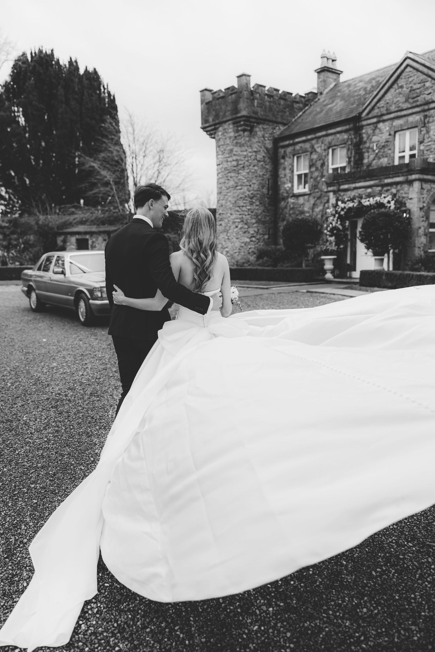 Black and white photo of bride and groom embracing outside a stone manor, bride's long train flowing behind her.