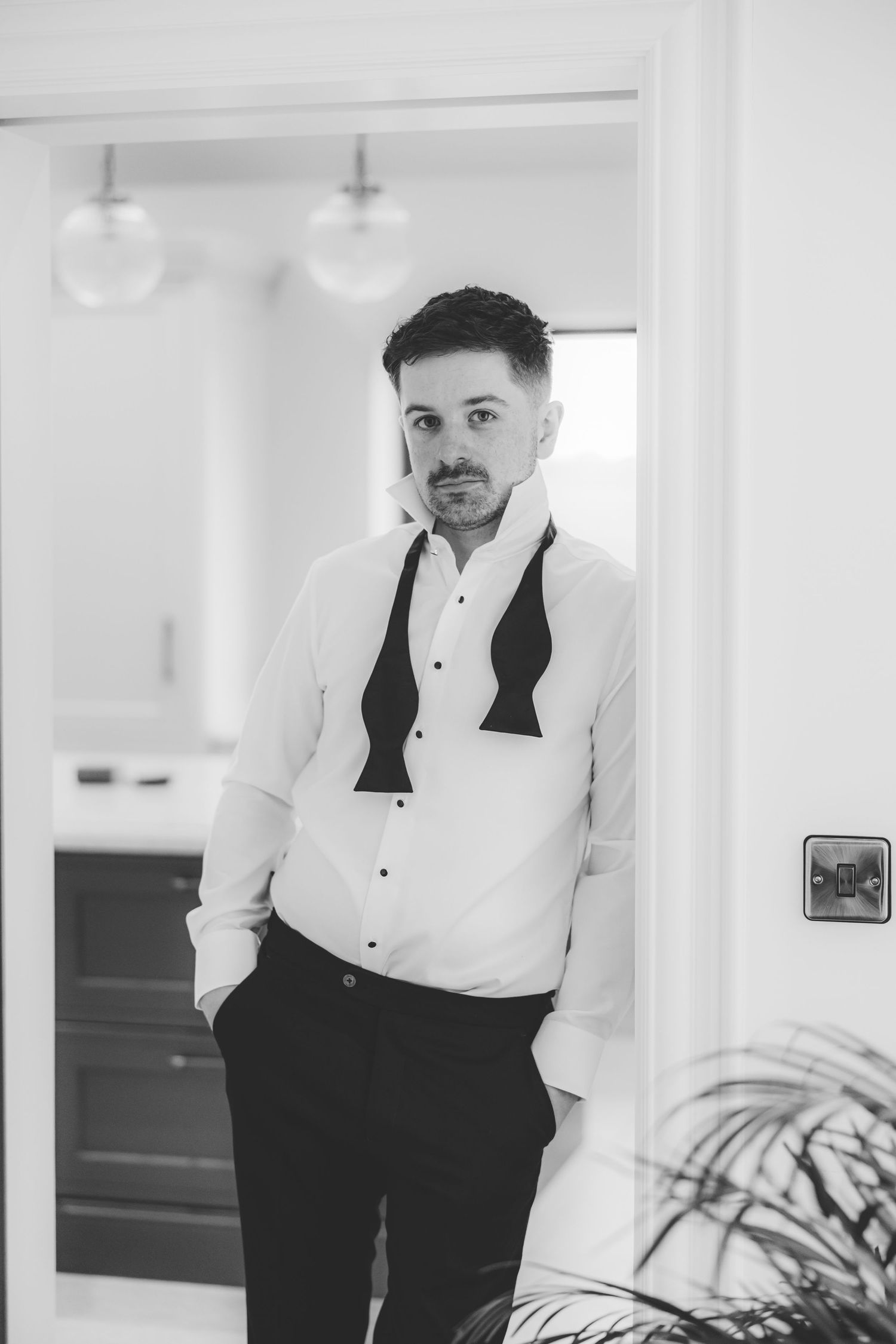 Black and white portrait of a well-dressed man in a tuxedo shirt and bow tie leaning against a doorframe.