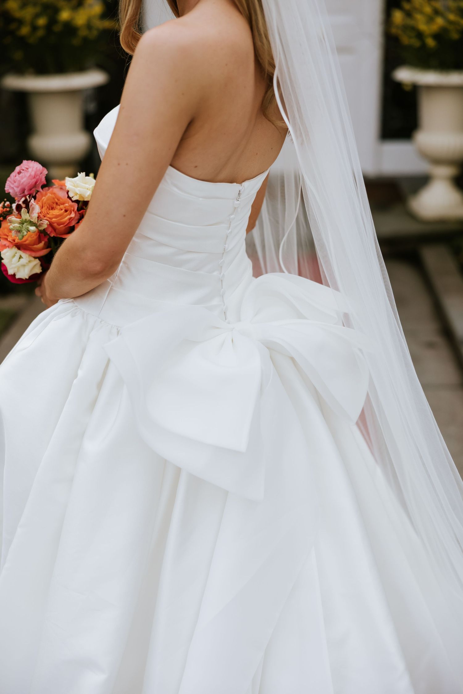Bride in strapless white ball gown with large bow and veil, holding colorful bouquet, viewed from behind.