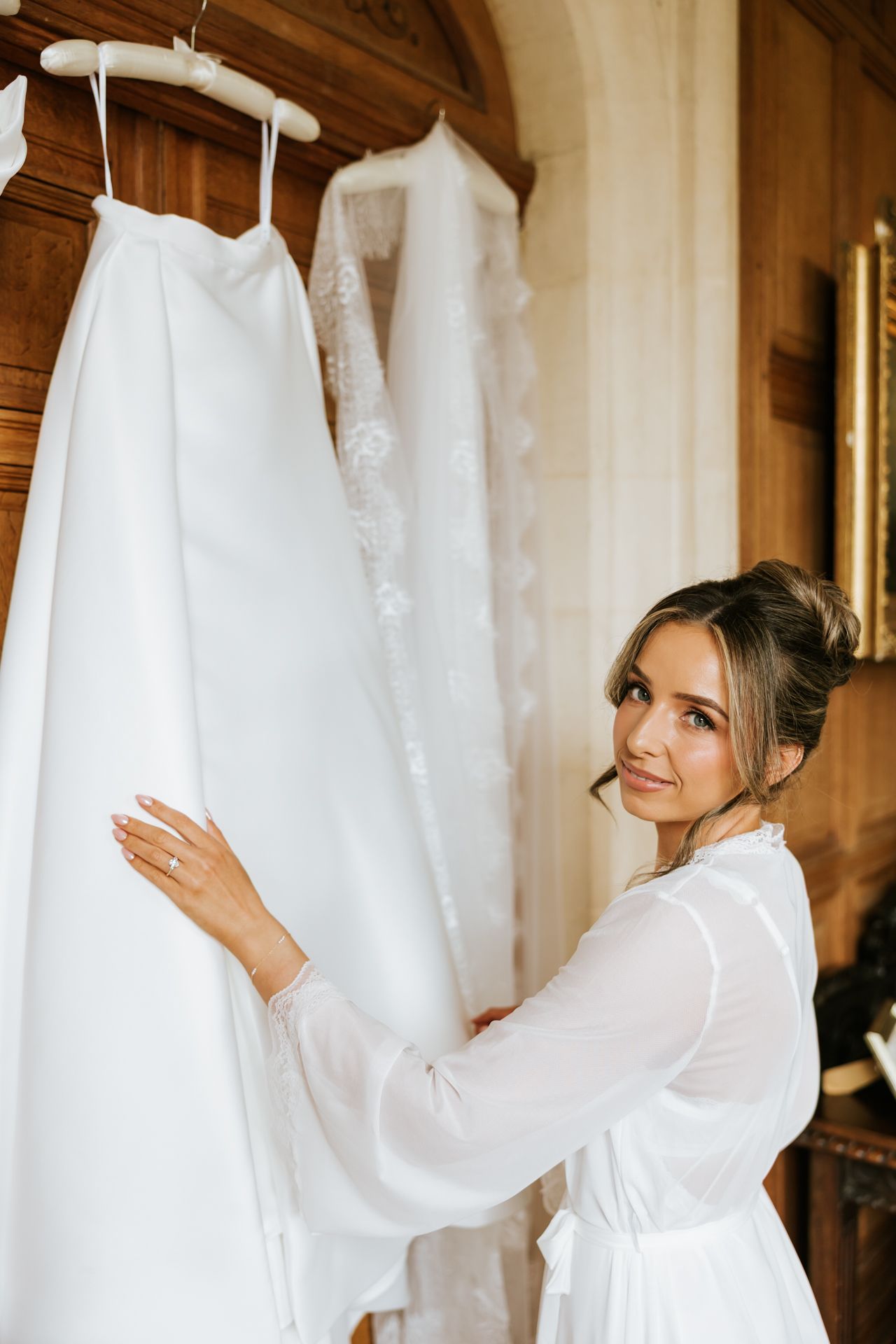 Bride in white robe admiring her wedding dress and veil hanging in an elegant wooden room.