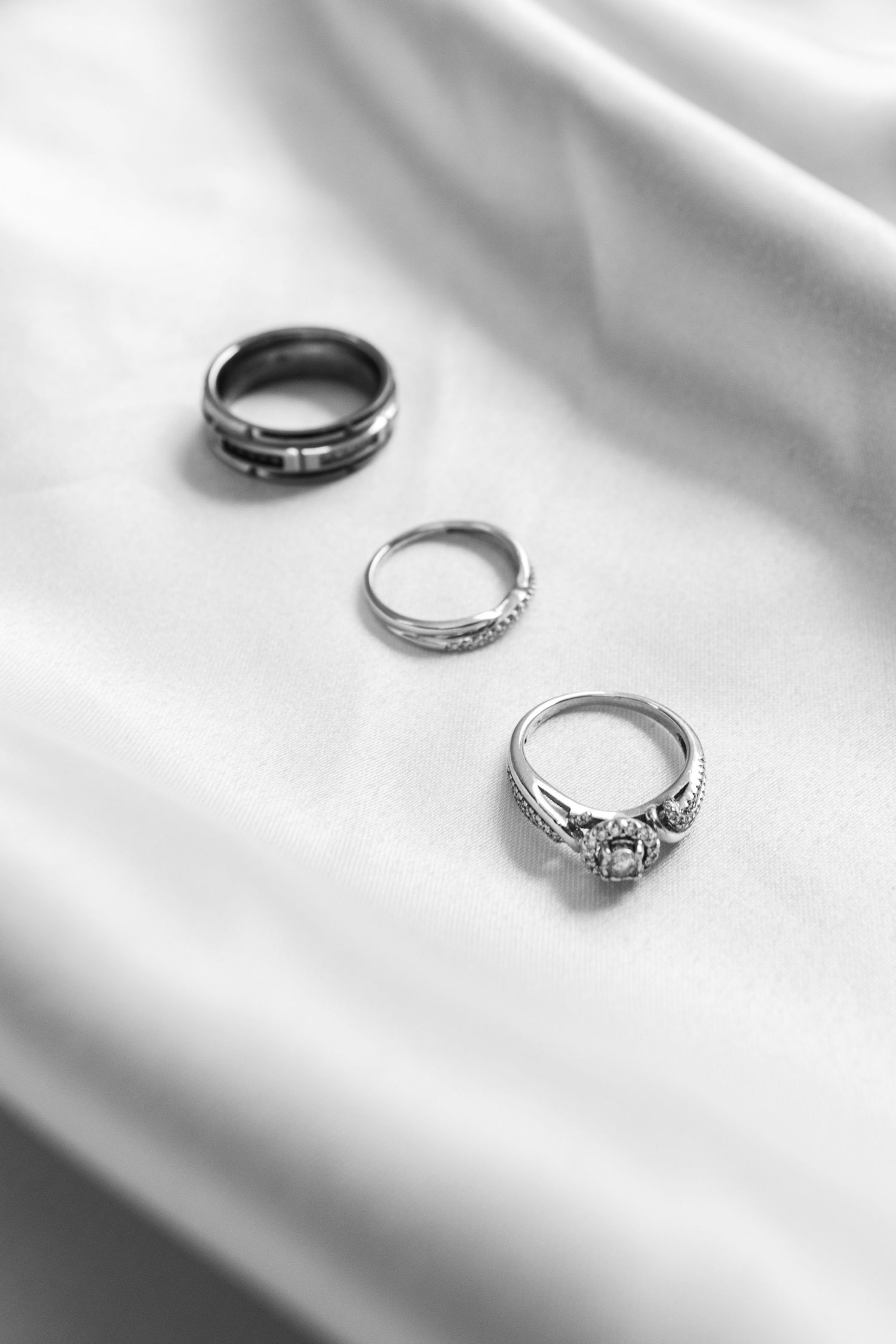 Three wedding rings arranged on white silk fabric, including a diamond engagement ring and two wedding bands in black and white.