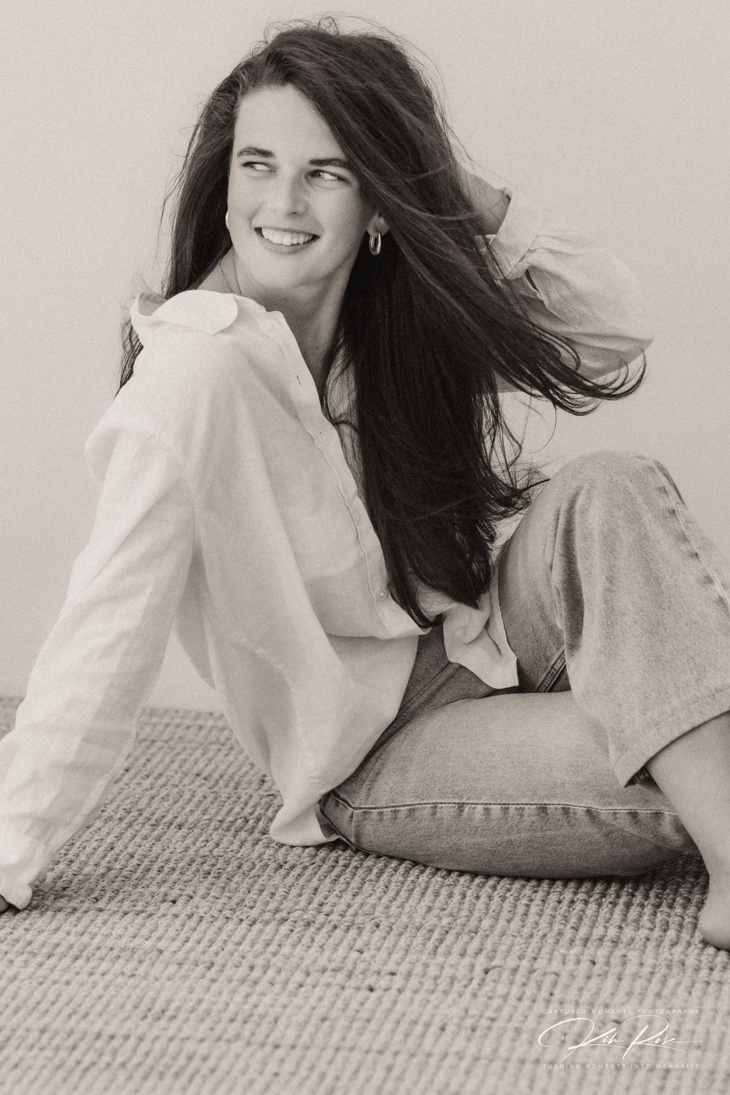Black and white photo of a smiling young woman with long dark hair sitting on a rug in a white shirt and jeans.