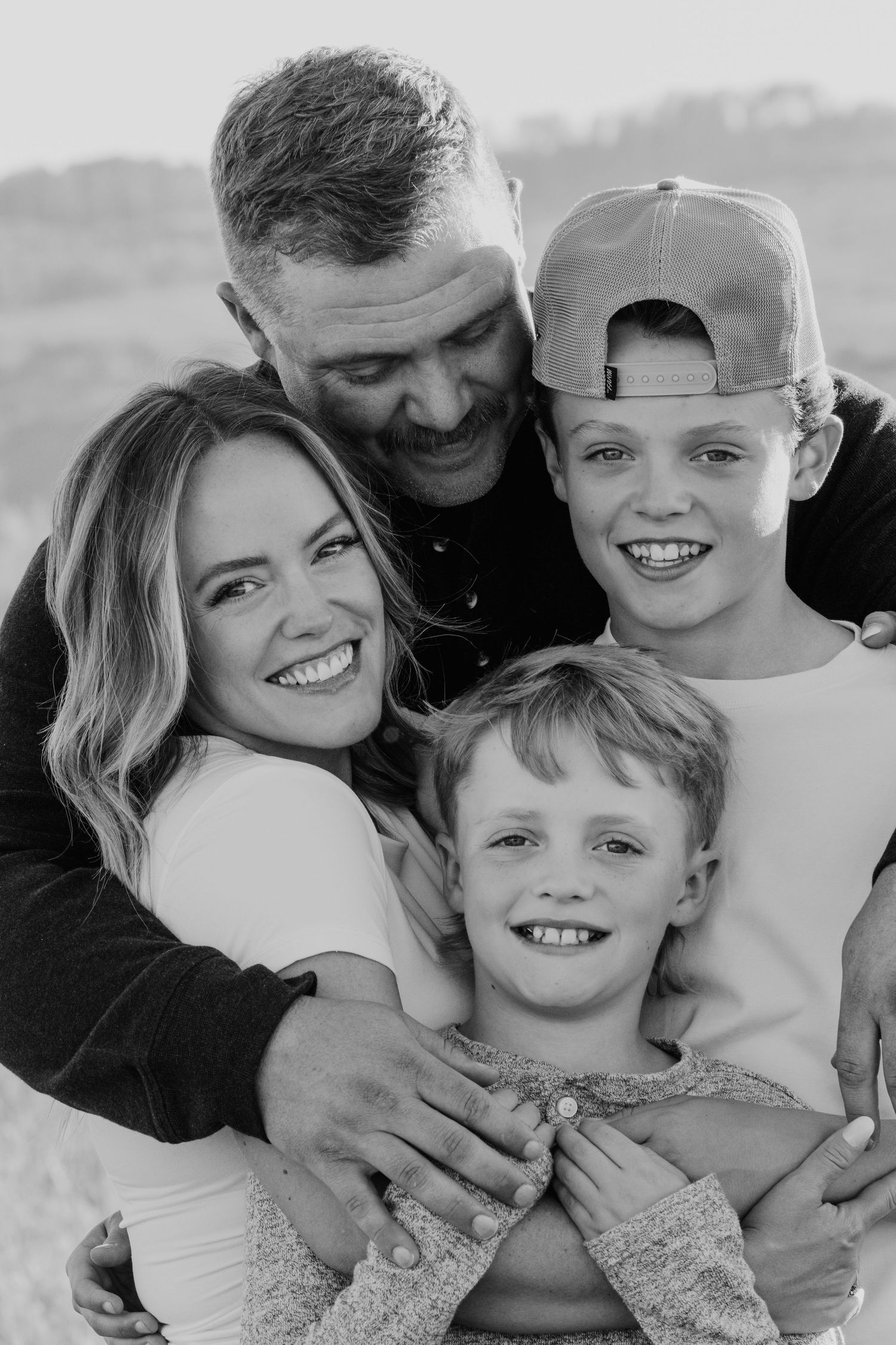Black and white family portrait of parents and two young boys smiling and embracing outdoors.