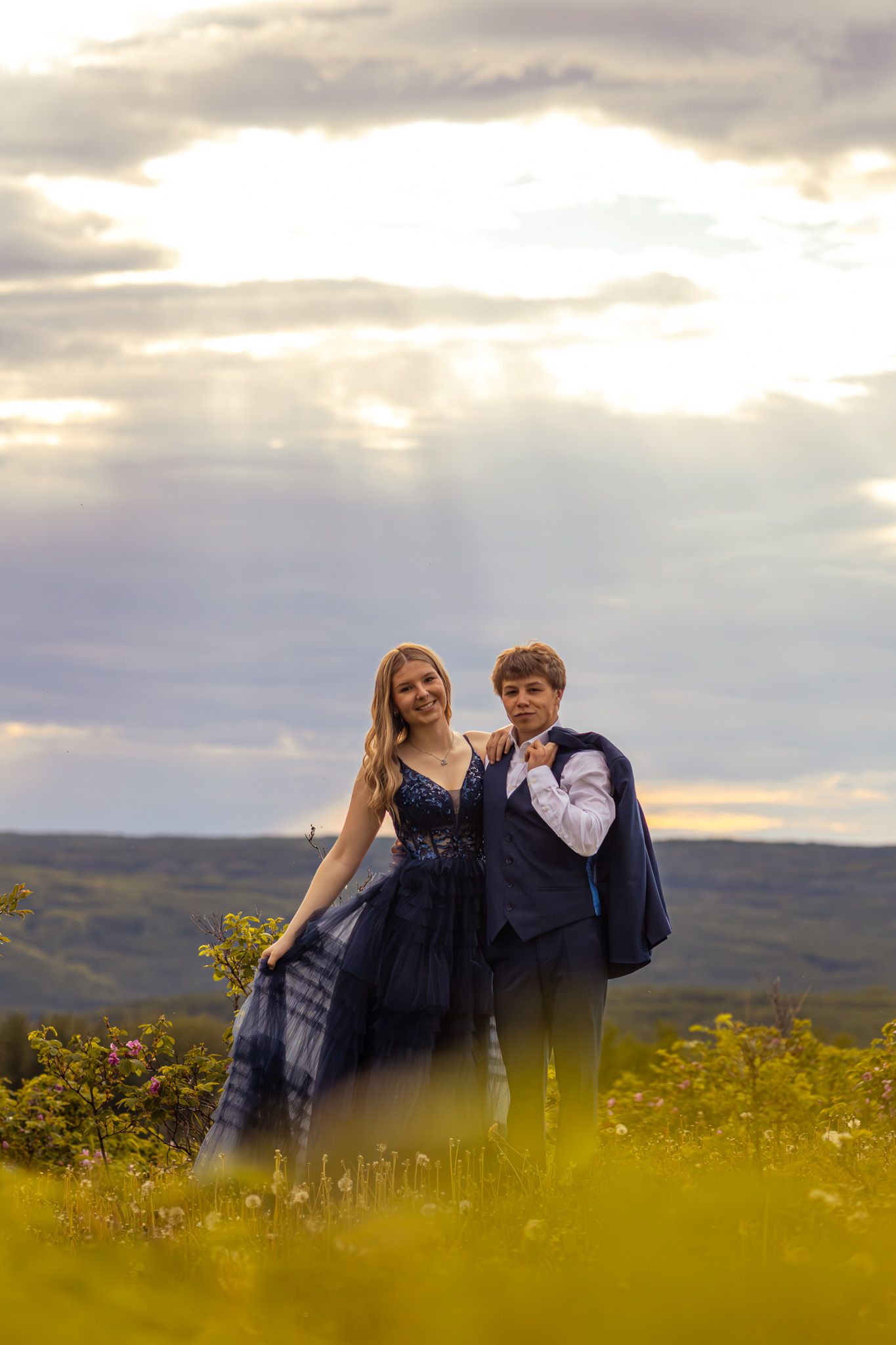 A young couple in matching navy blue formal attire poses outdoors at sunset with rolling hills in the background.
