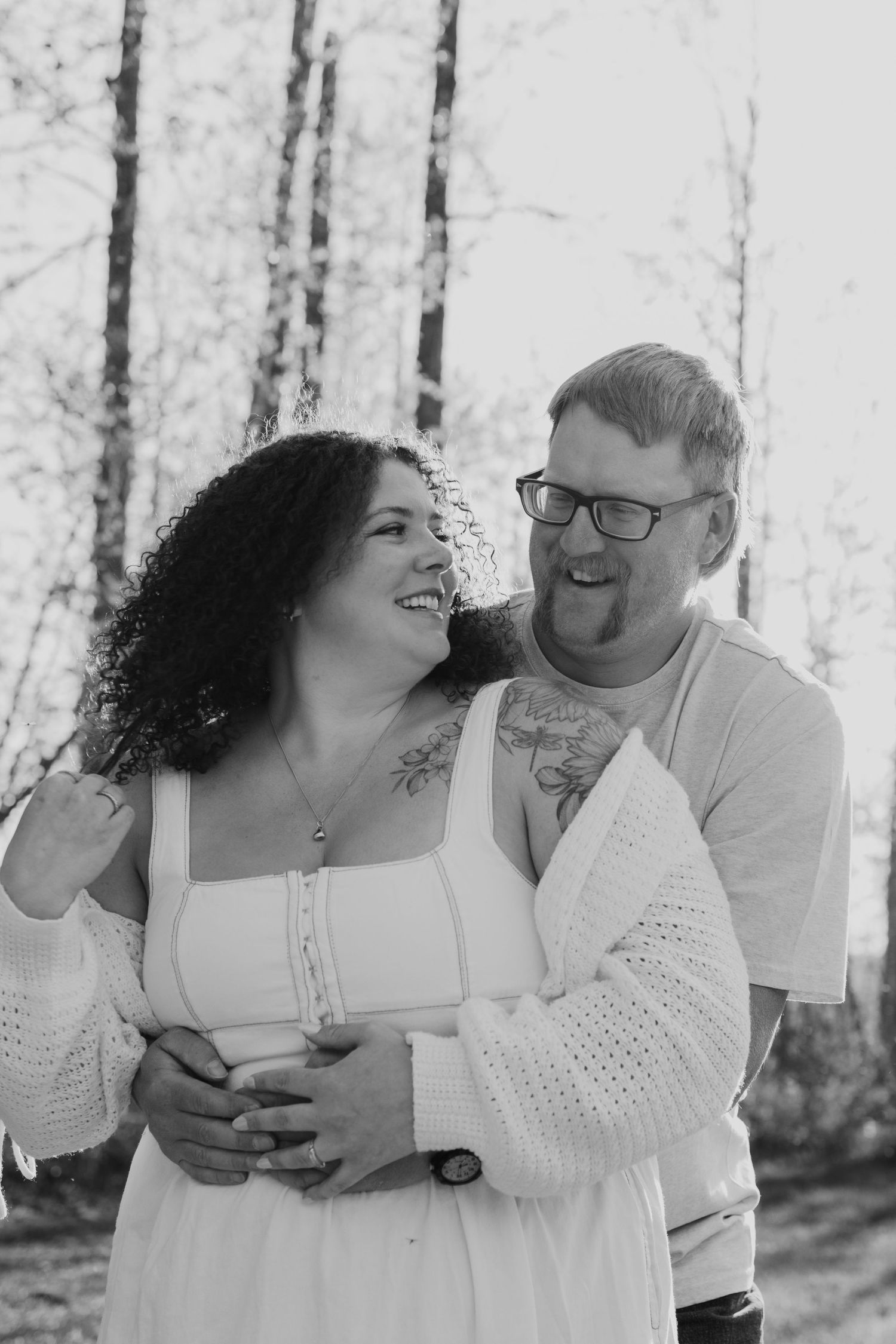 Black and white photo of a laughing couple embracing outdoors, woman in white dress with tattoos, man in glasses hugging her.