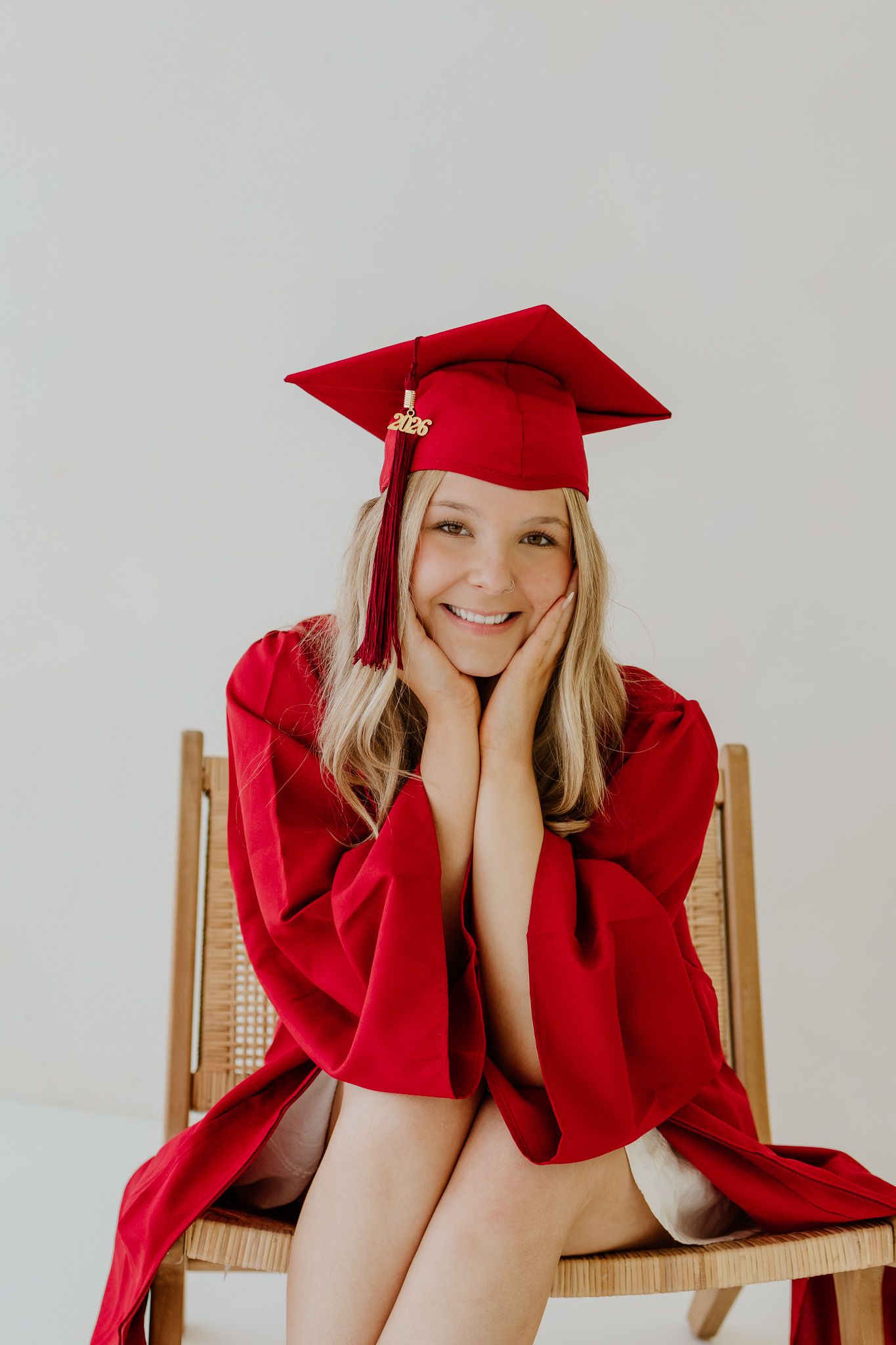 Smiling graduate in red cap and gown sitting on a wooden chair, posing with hands on cheeks for graduation photos.