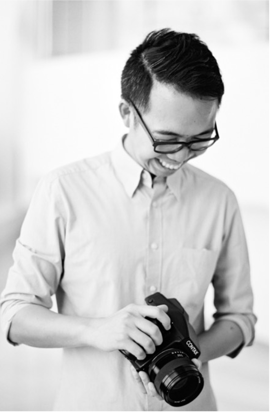 Smiling young man with glasses holding a Canon camera, wearing a light button-up shirt in a black and white photo.