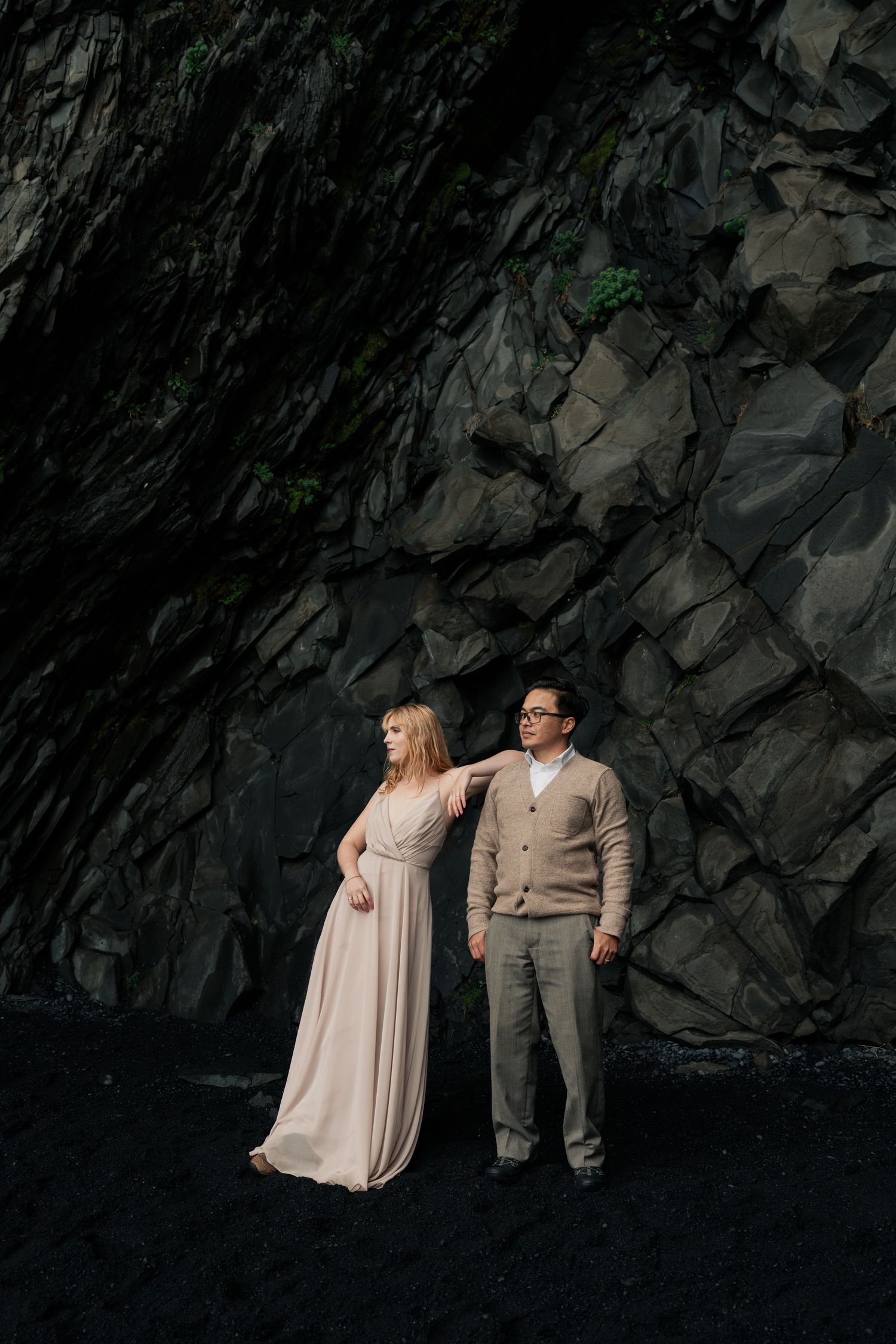 Couple posing against dark volcanic rock wall on black sand beach, woman in blush gown, man in gray suit.