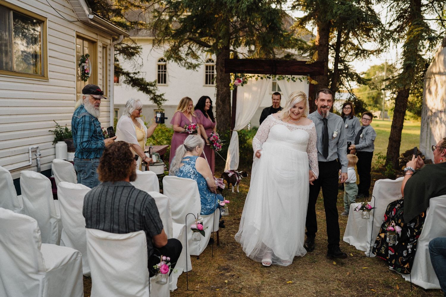 Bride and groom walking down outdoor wedding aisle surrounded by guests and white chairs.