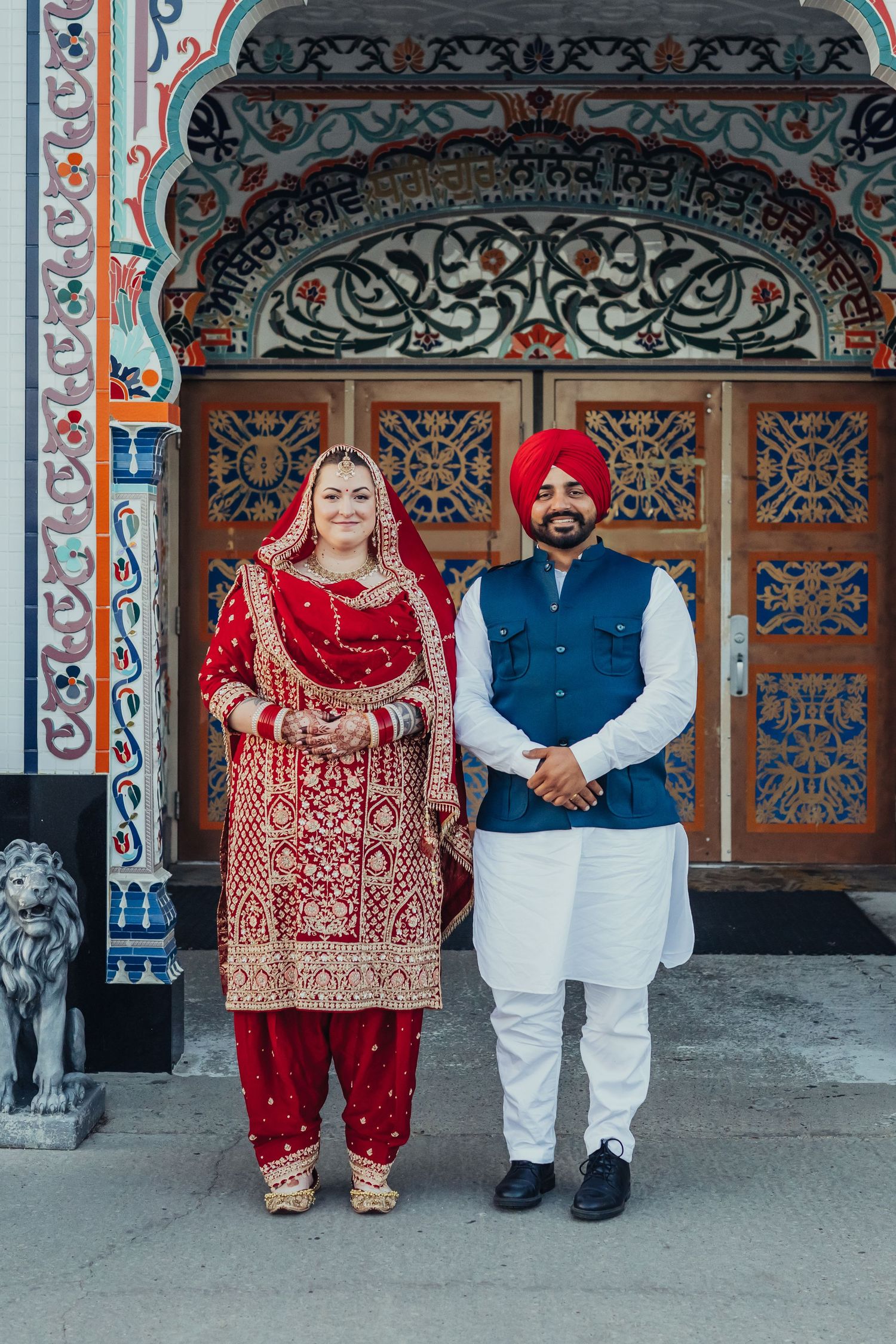 Sikh couple in traditional red and blue wedding attire posing before ornate wooden doors with colorful architecture.