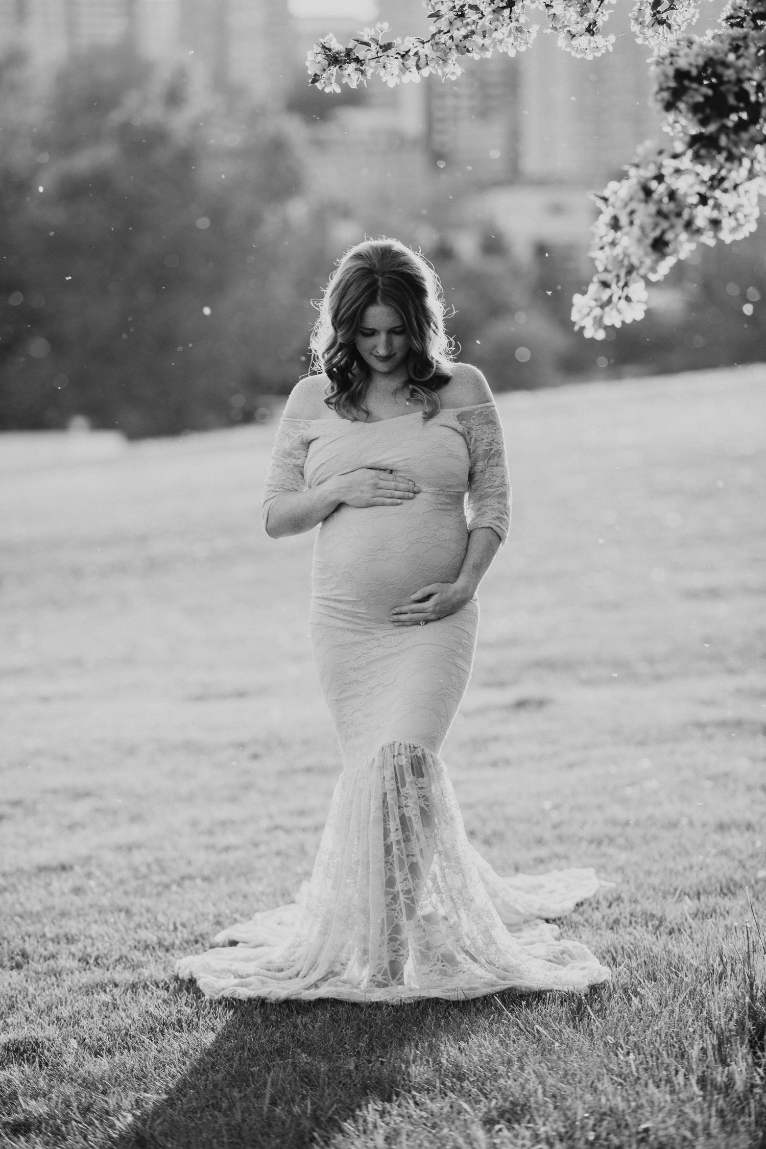 Black and white maternity photo of pregnant woman in lace gown holding belly under flowering tree in park.