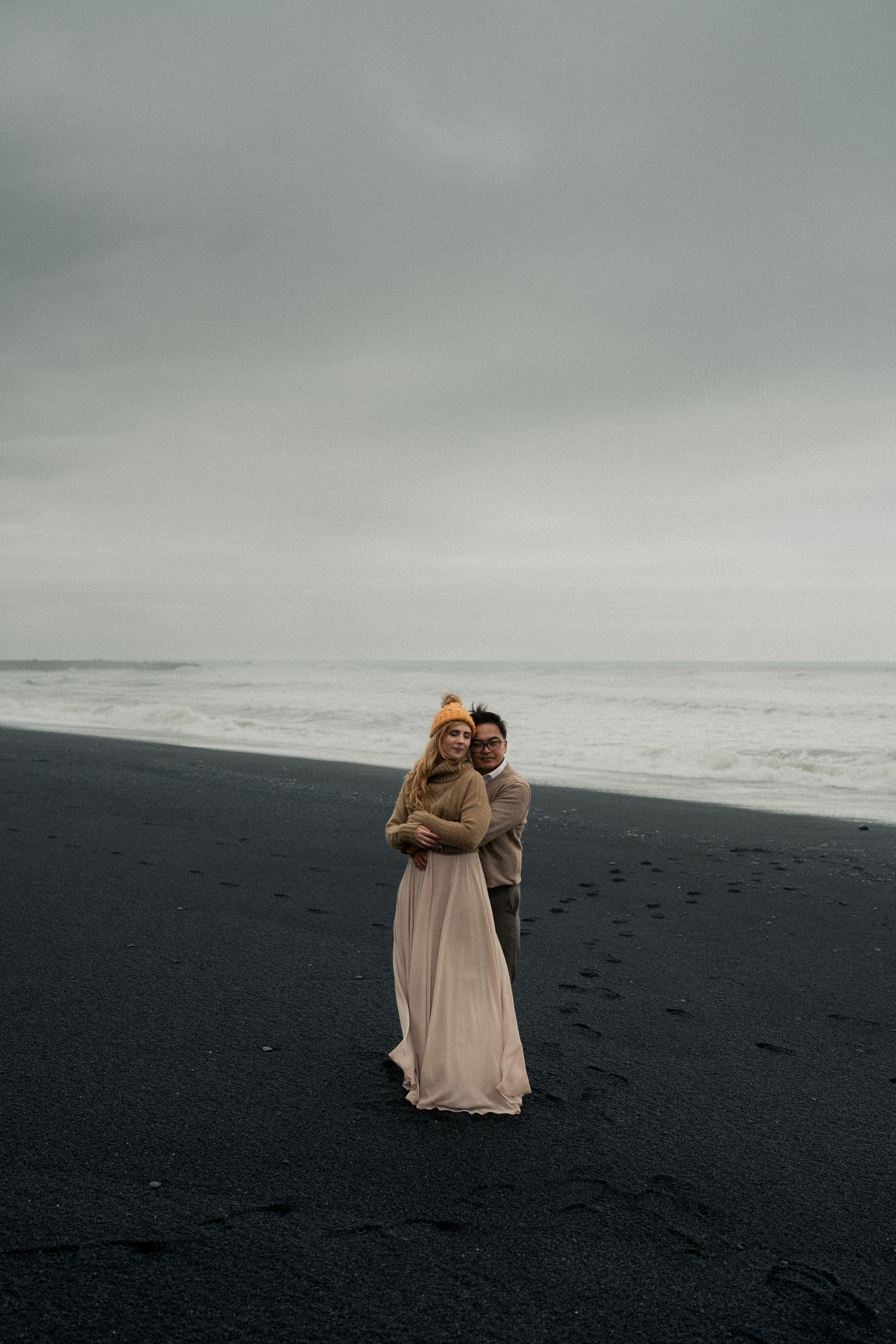 Couple embracing on a black sand beach in Iceland, wearing elegant attire, with moody overcast skies and ocean waves behind them.