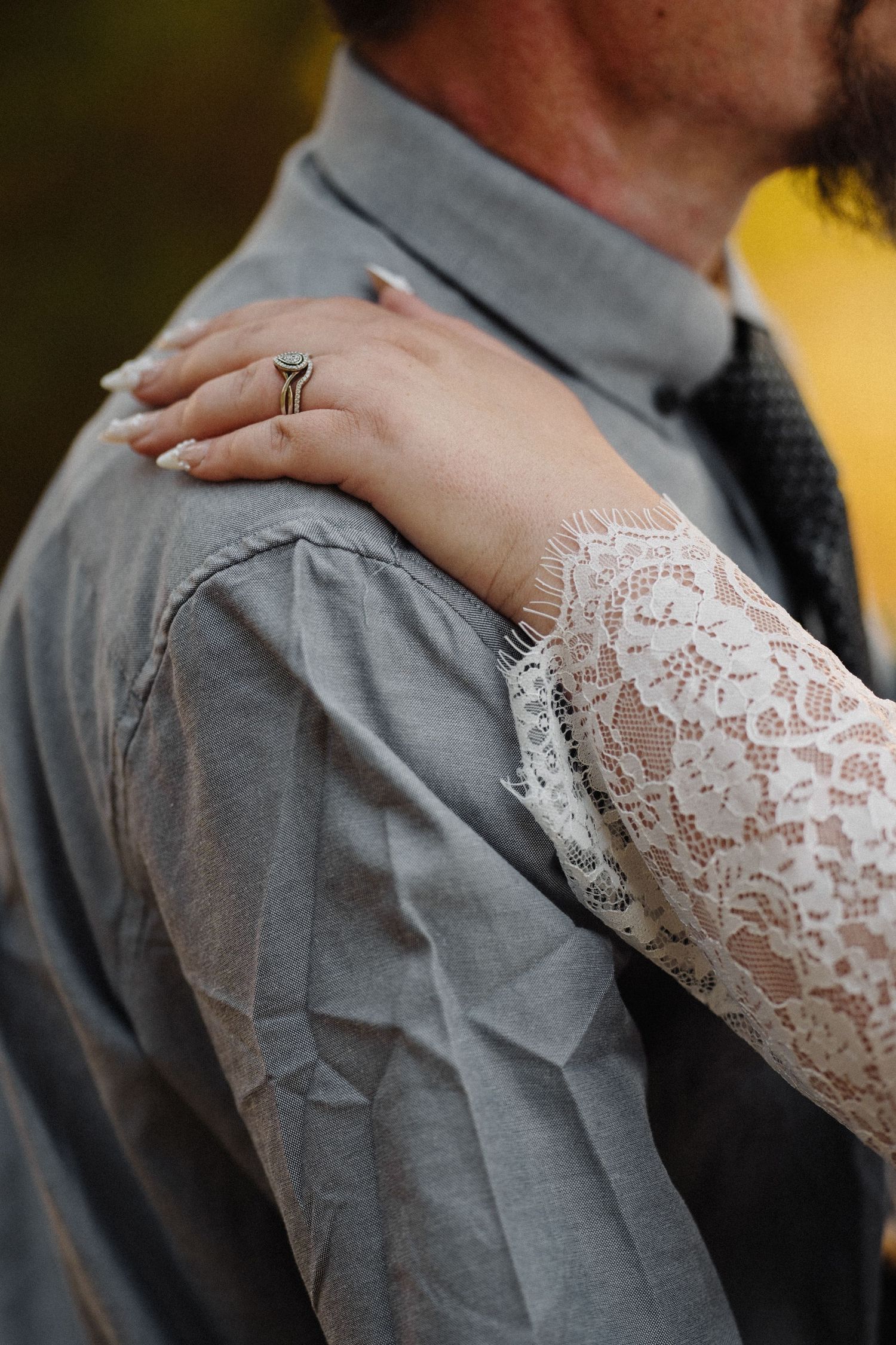 Close-up of a woman's hand with an engagement ring resting on a man's shoulder in a gray shirt.