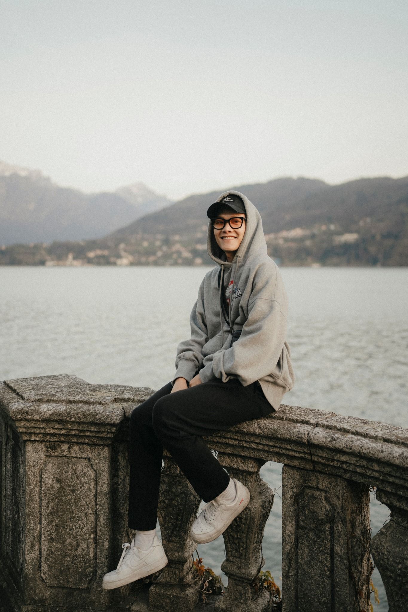 Person sitting on stone railing overlooking a mountain lake wearing casual grey hoodie and sneakers.
