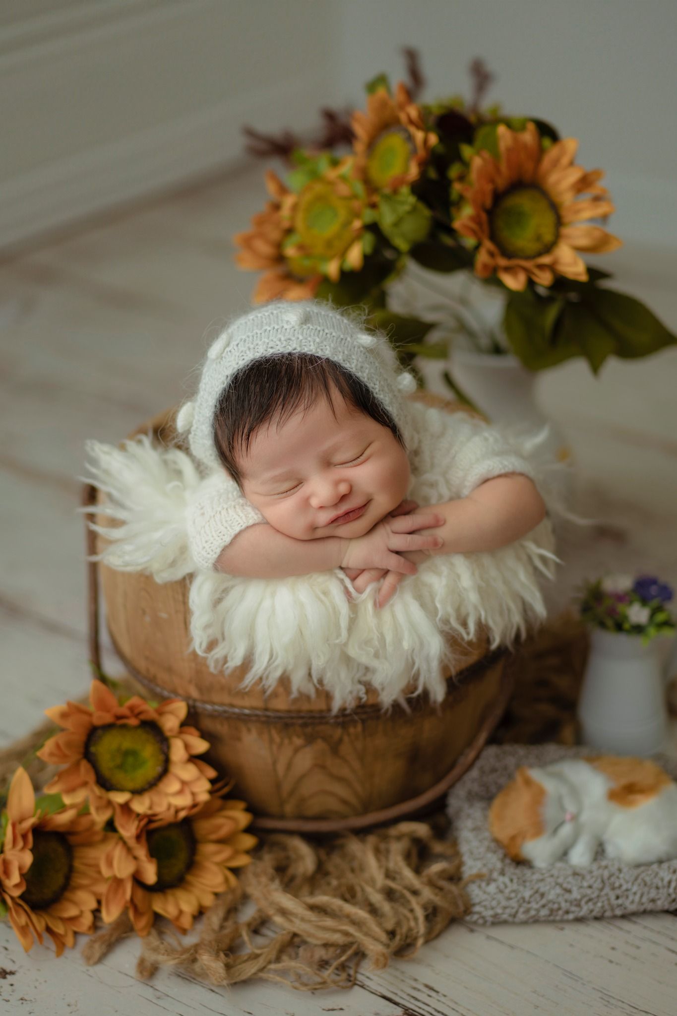 A peaceful sleeping baby in white knit bonnet nestled in fuzzy basket surrounded by sunflowers.