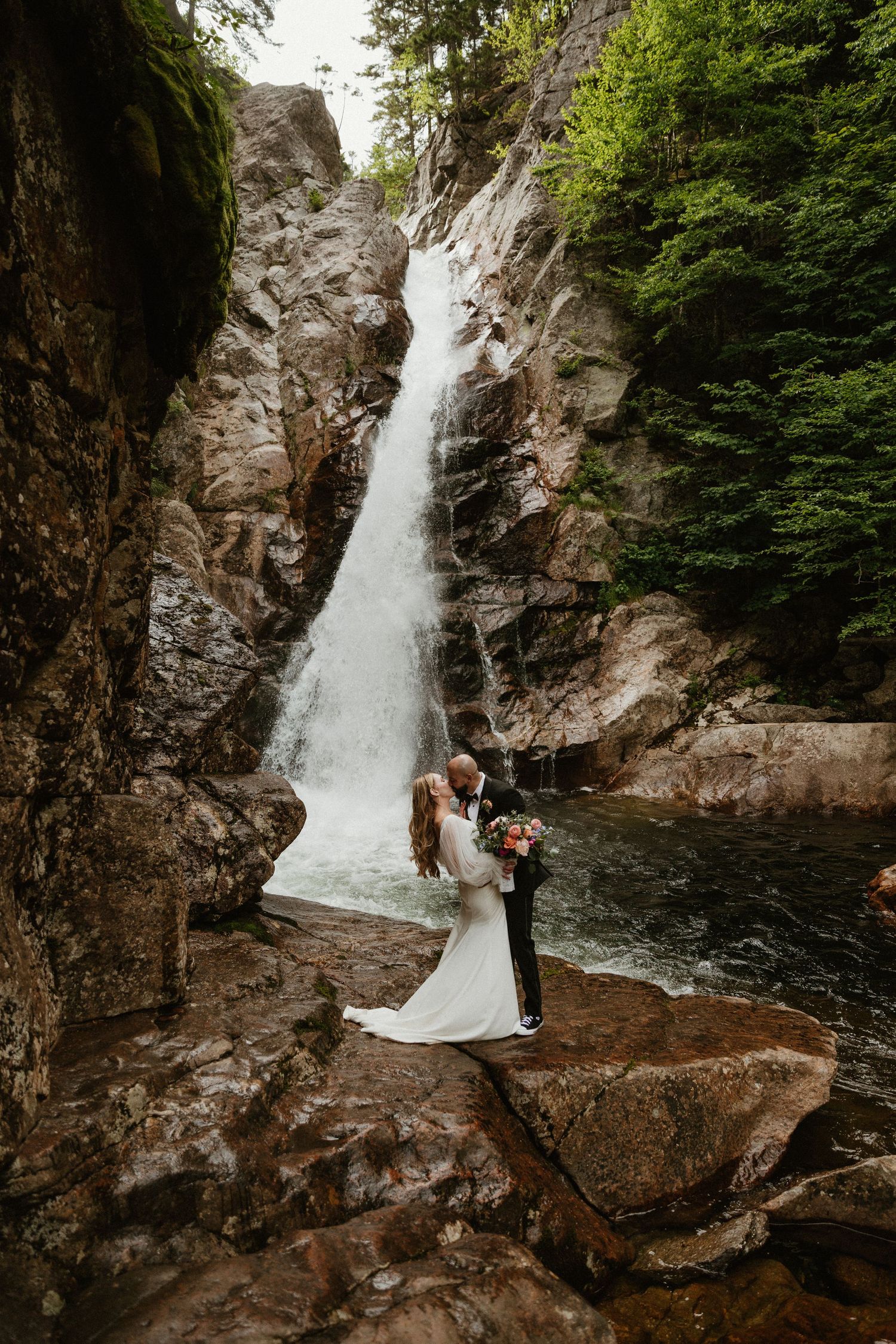 A dramatic waterfall creates a stunning backdrop for an intimate wedding moment on rocky terrain.