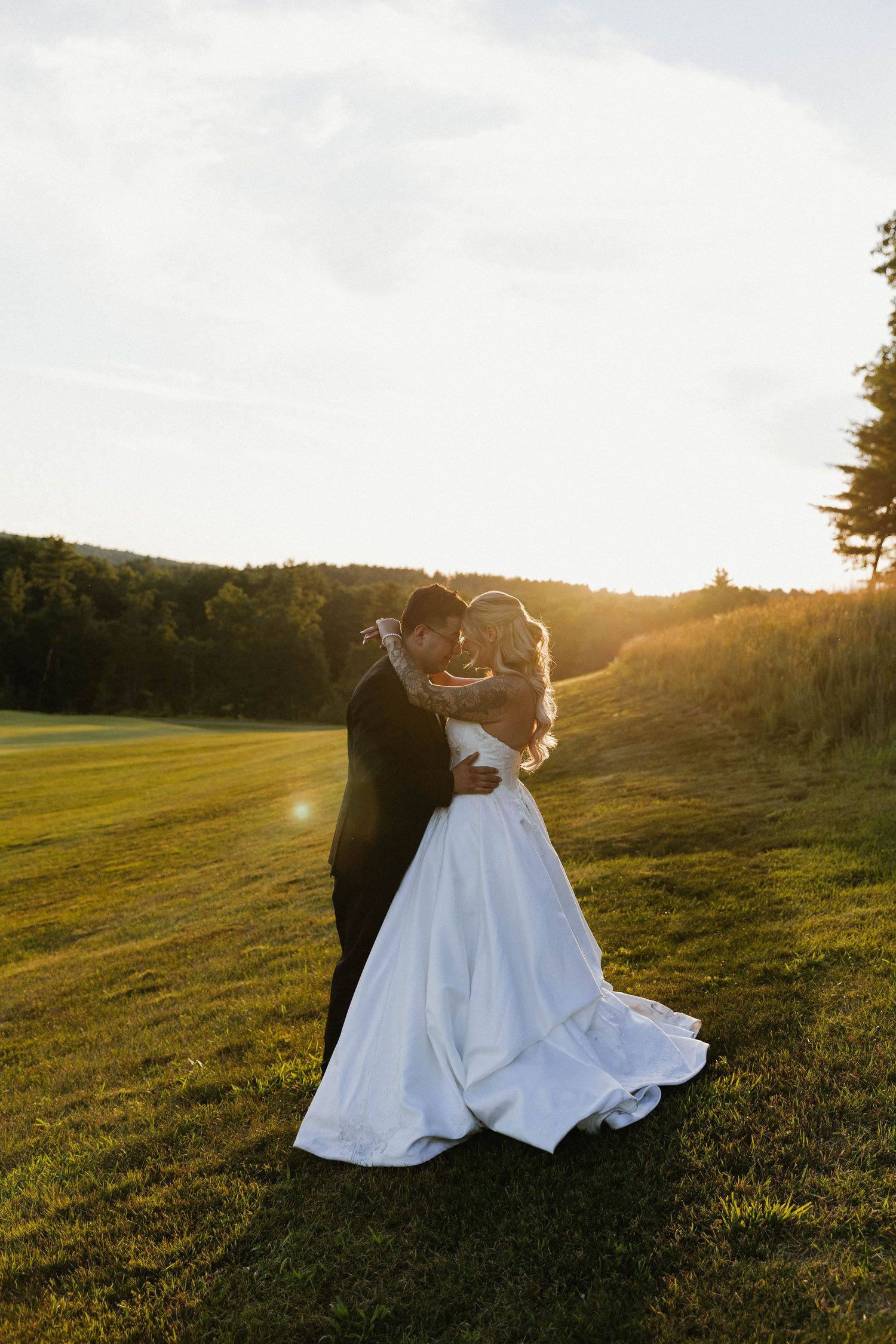 Bride and groom embracing at golden hour sunset on lush green field, romantic outdoor wedding portrait.