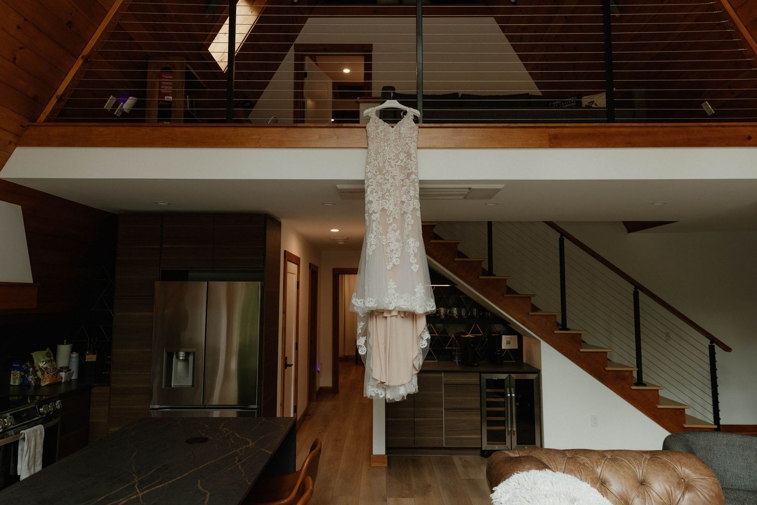 A elegant lace wedding dress hanging on a hanger in a modern rustic home with wood ceilings and staircase.