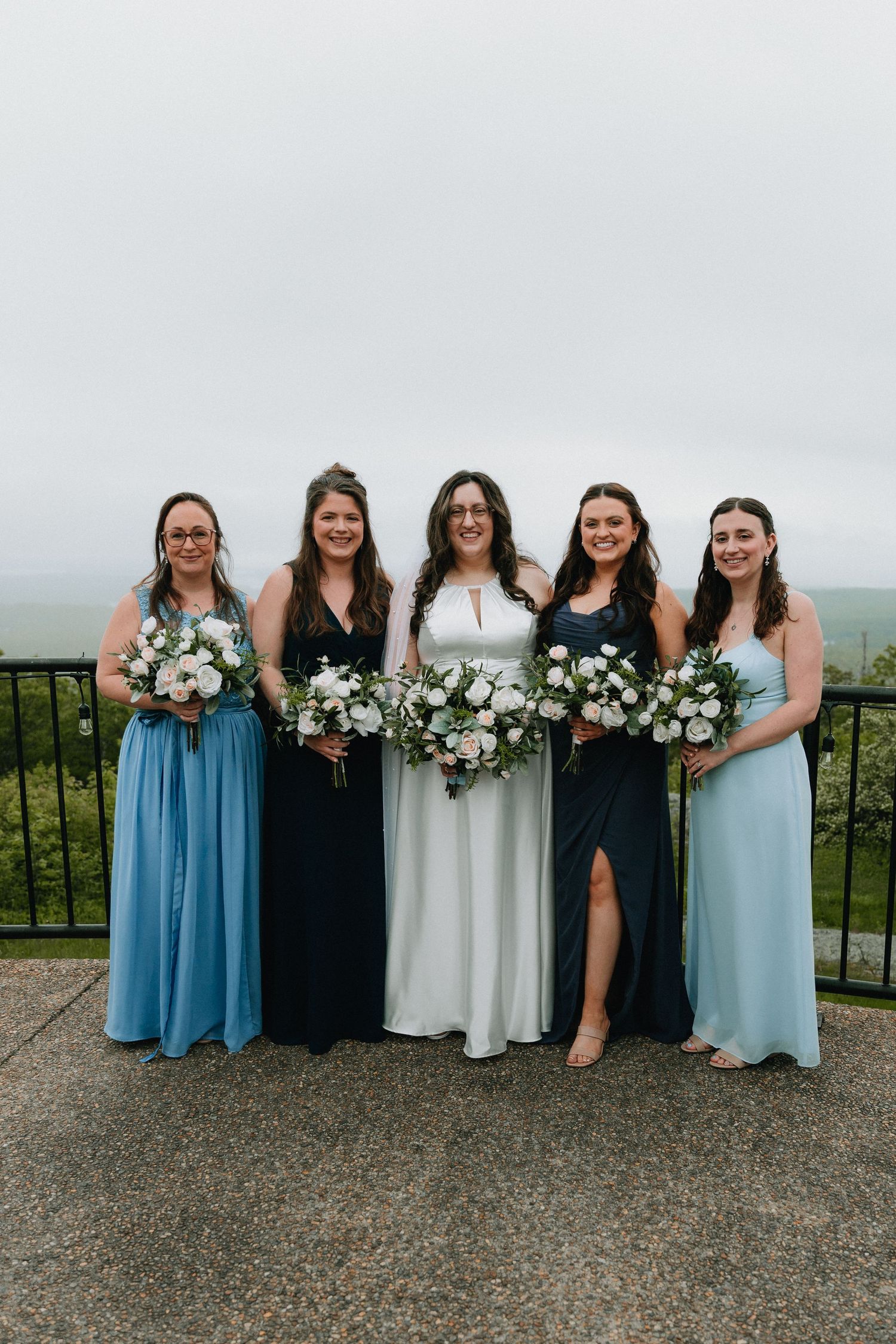 Wedding party in alternating blue and navy dresses posing with bouquets on stone patio.