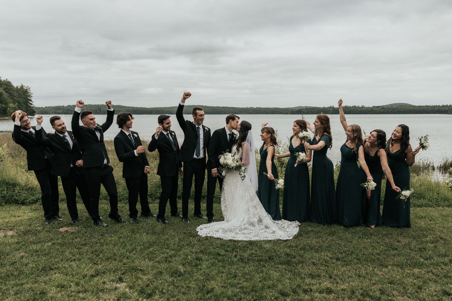 Wedding party lined up for group photo by waterfront on overcast day.