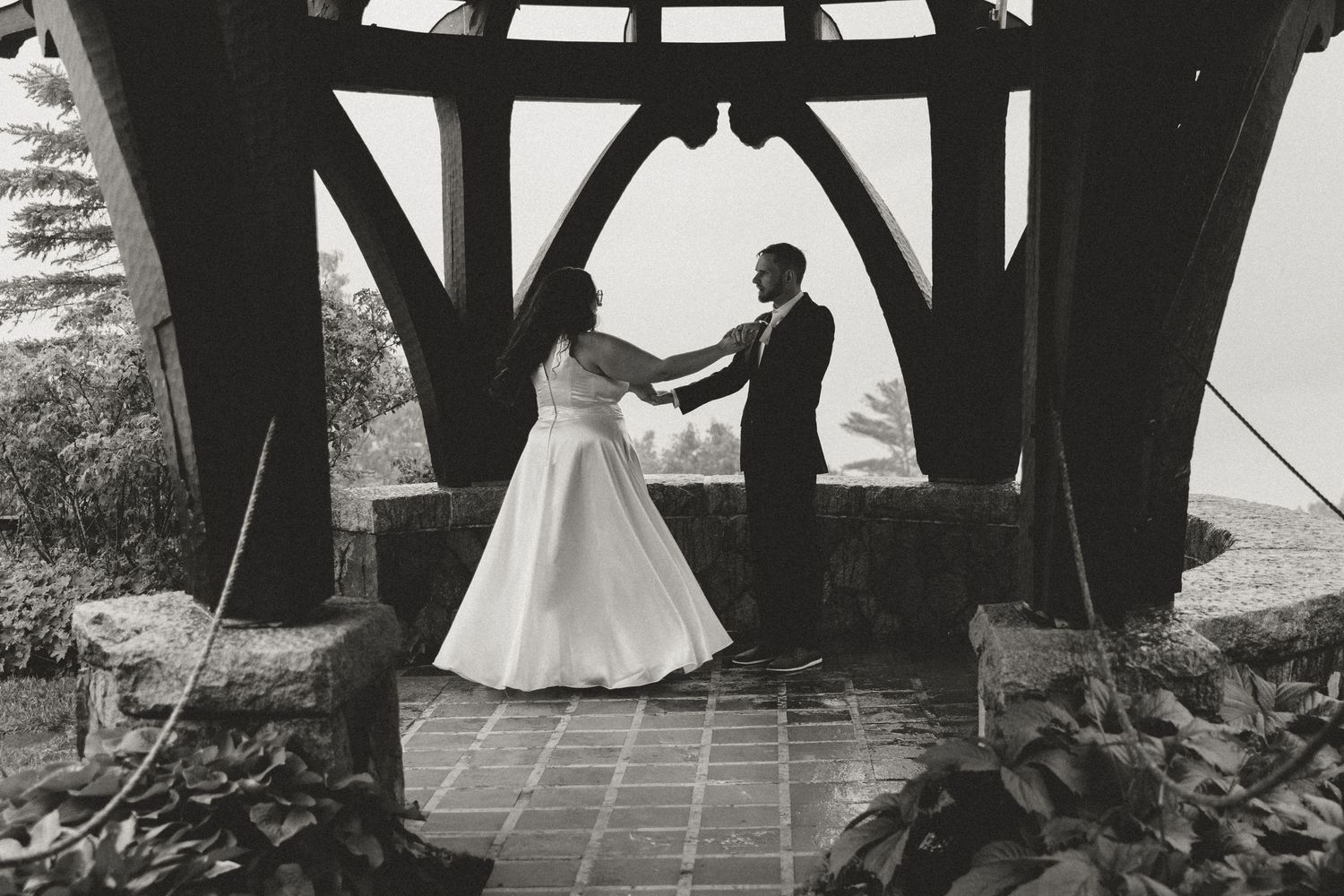 Black and white photo of wedding ceremony under rustic wooden gazebo.