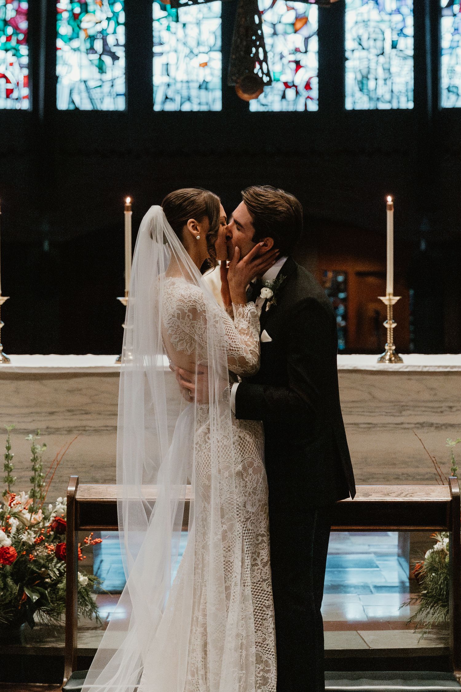 Romantic wedding kiss in front of stained glass windows with glowing candles.