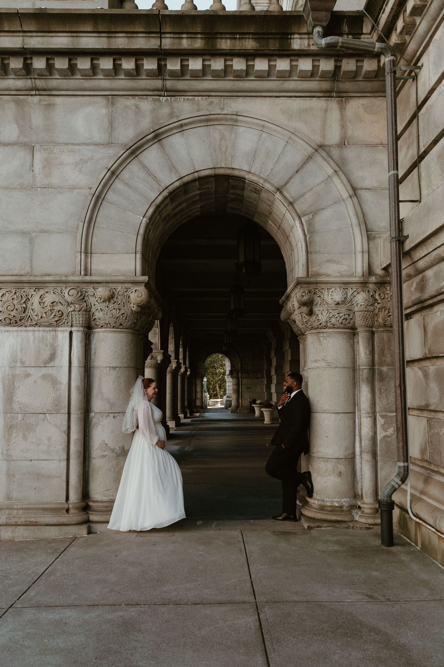 A wedding couple stands beneath ornate stone archways in a classical architectural setting.