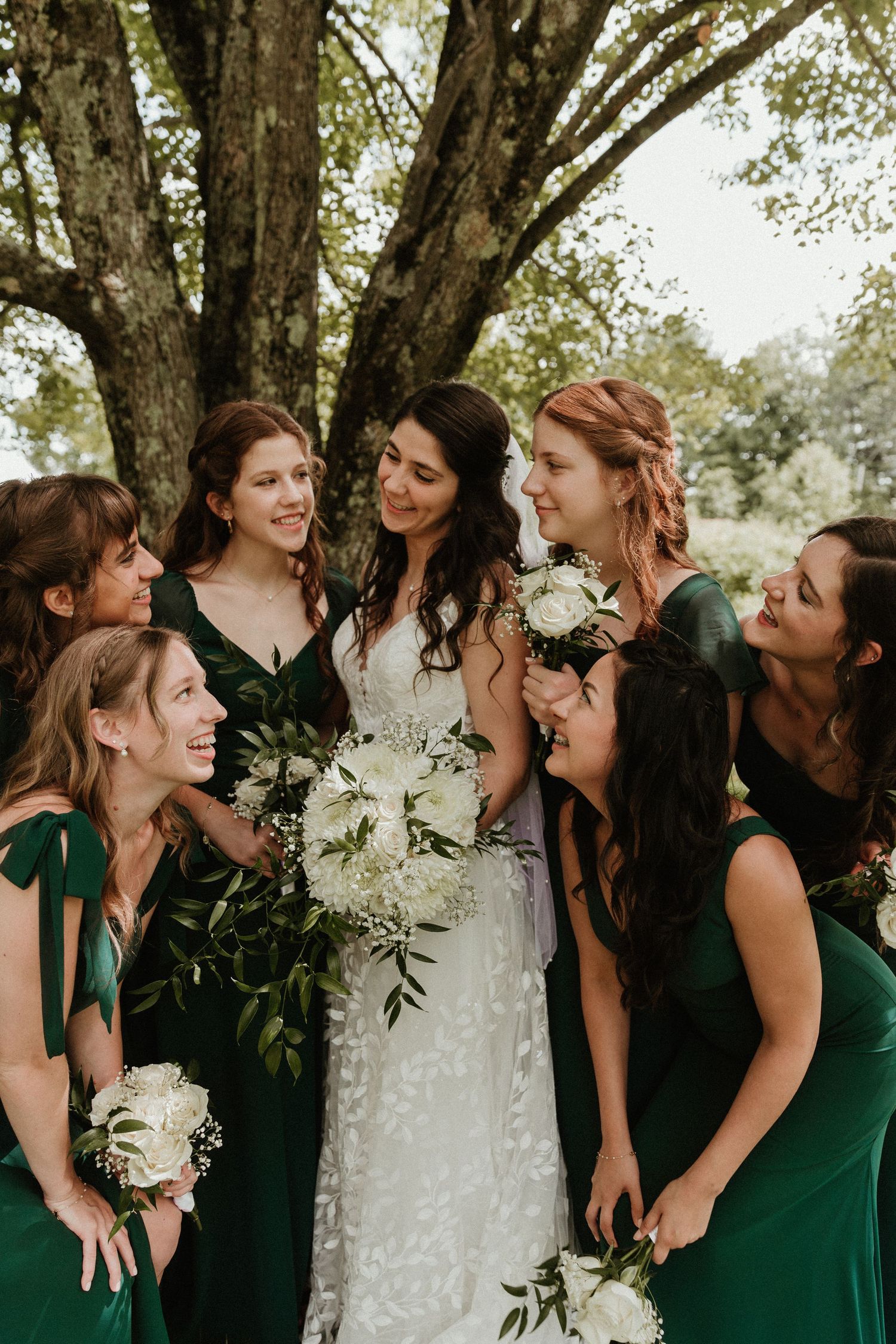 Bridesmaids in elegant emerald green dresses gather around bride in white lace gown under trees.