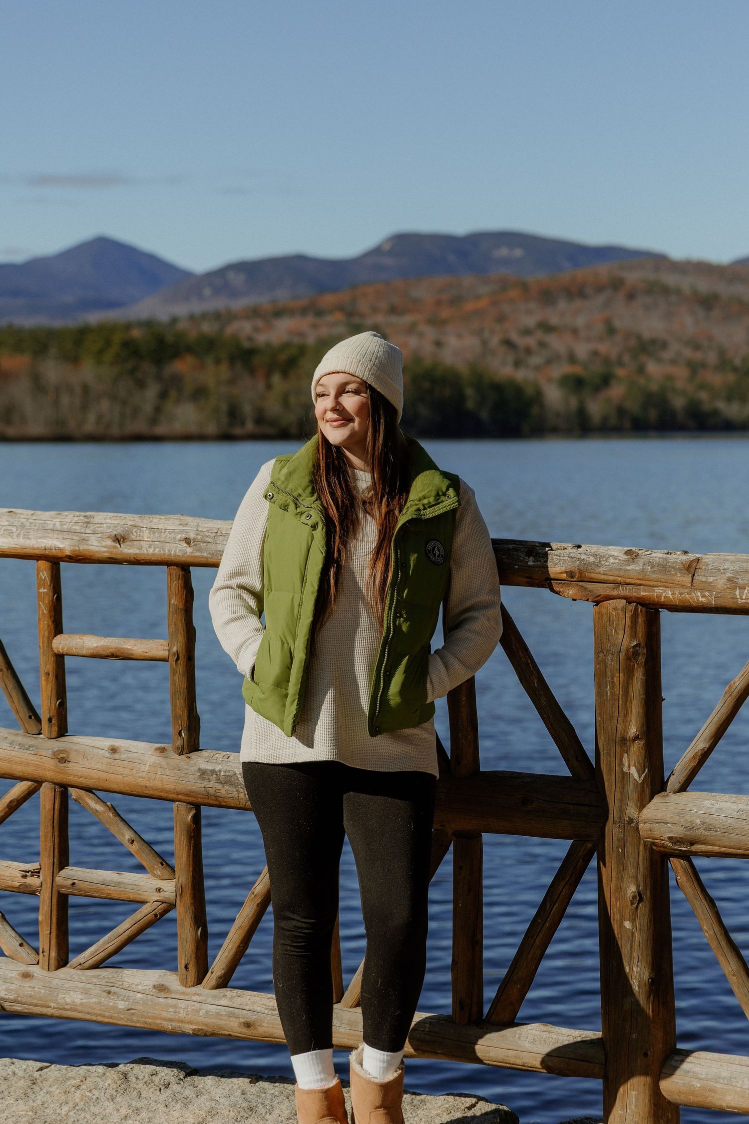 Person in winter attire leans on rustic wooden railing overlooking a serene mountain lake on an autumn day.