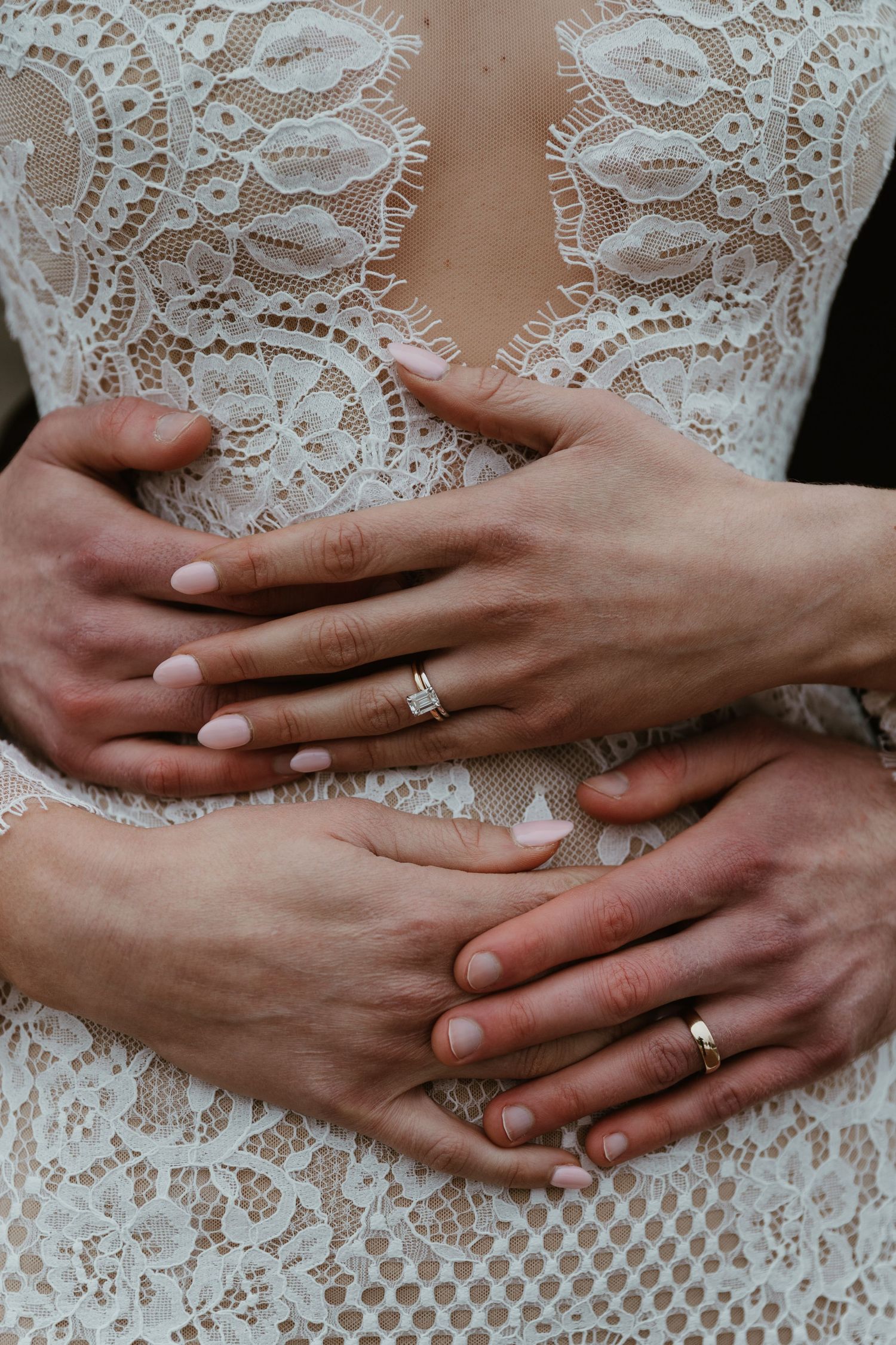 Close-up of hands with wedding rings against intricate white lace wedding dress detail.