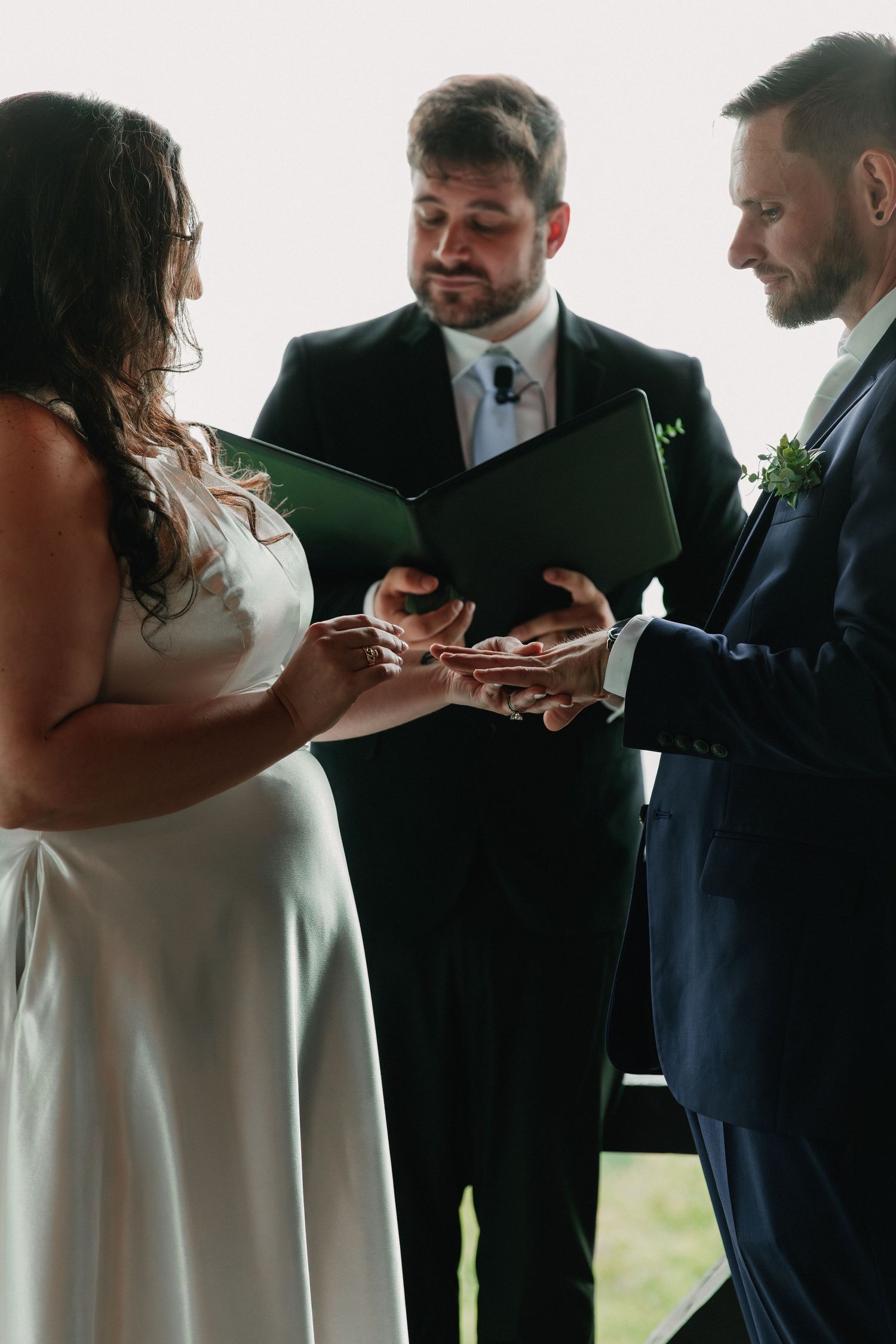 Wedding ceremony participants in formal attire exchanging rings by a window.