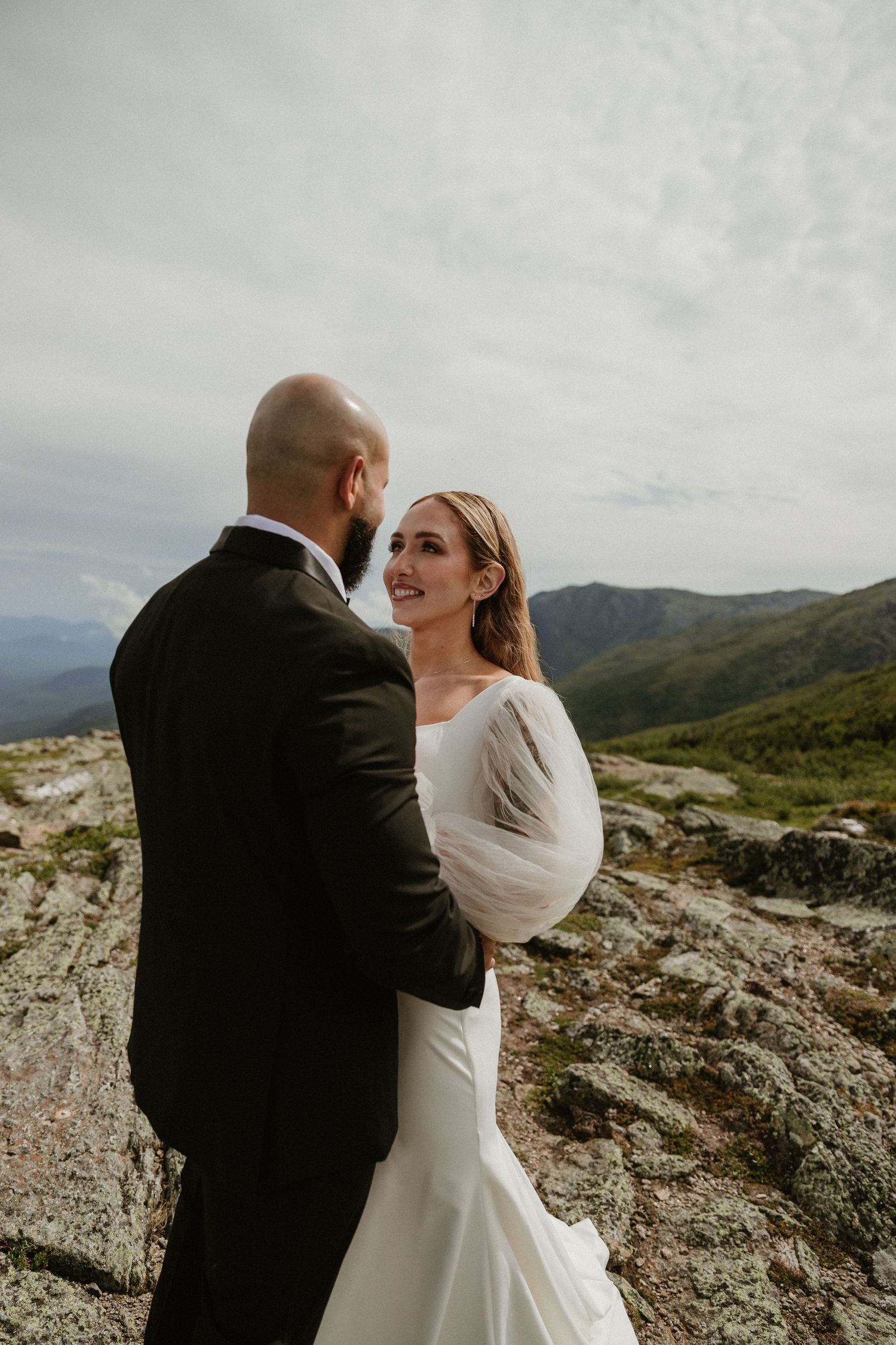 A couple in wedding attire shares a romantic moment on a mountain peak with cloudy skies.