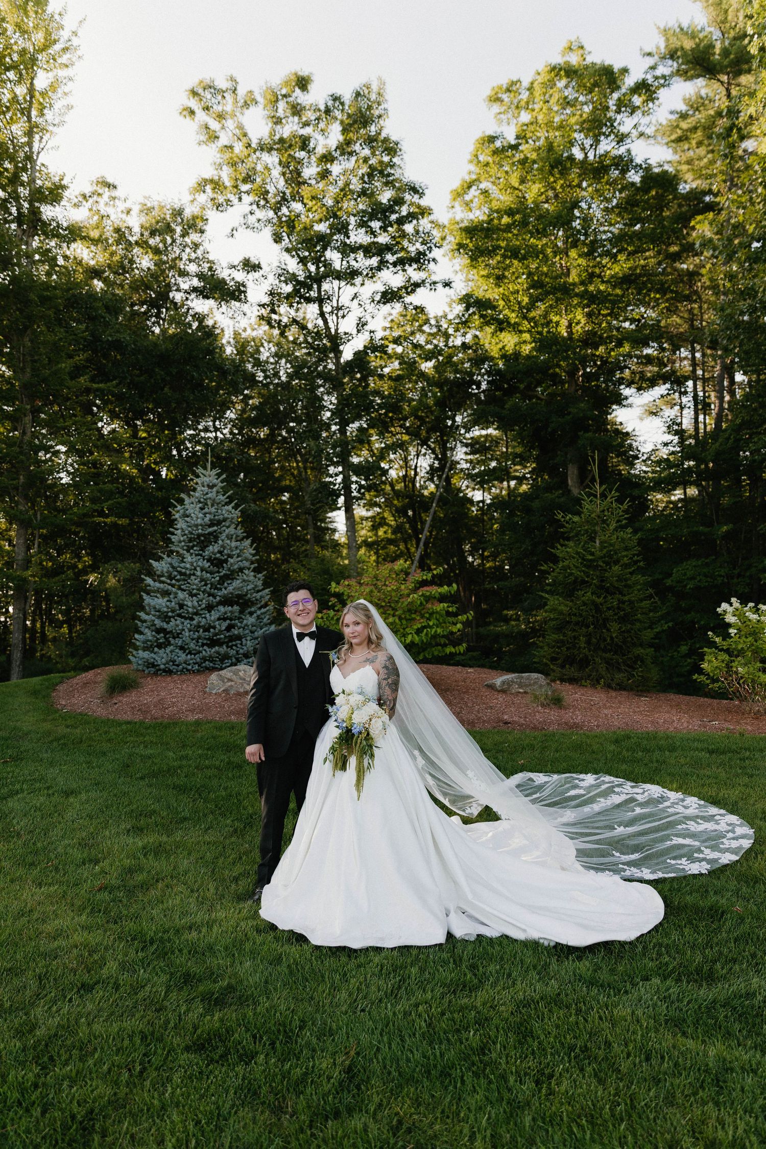 Bride in white ball gown with veil and groom in black tuxedo posing on green lawn surrounded by lush trees.