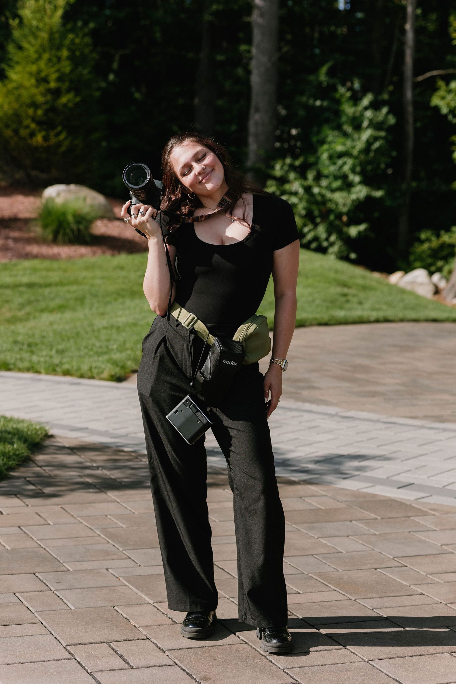 Young woman in black outfit holding a camera lens outdoors on a sunny day in a lush green park setting.