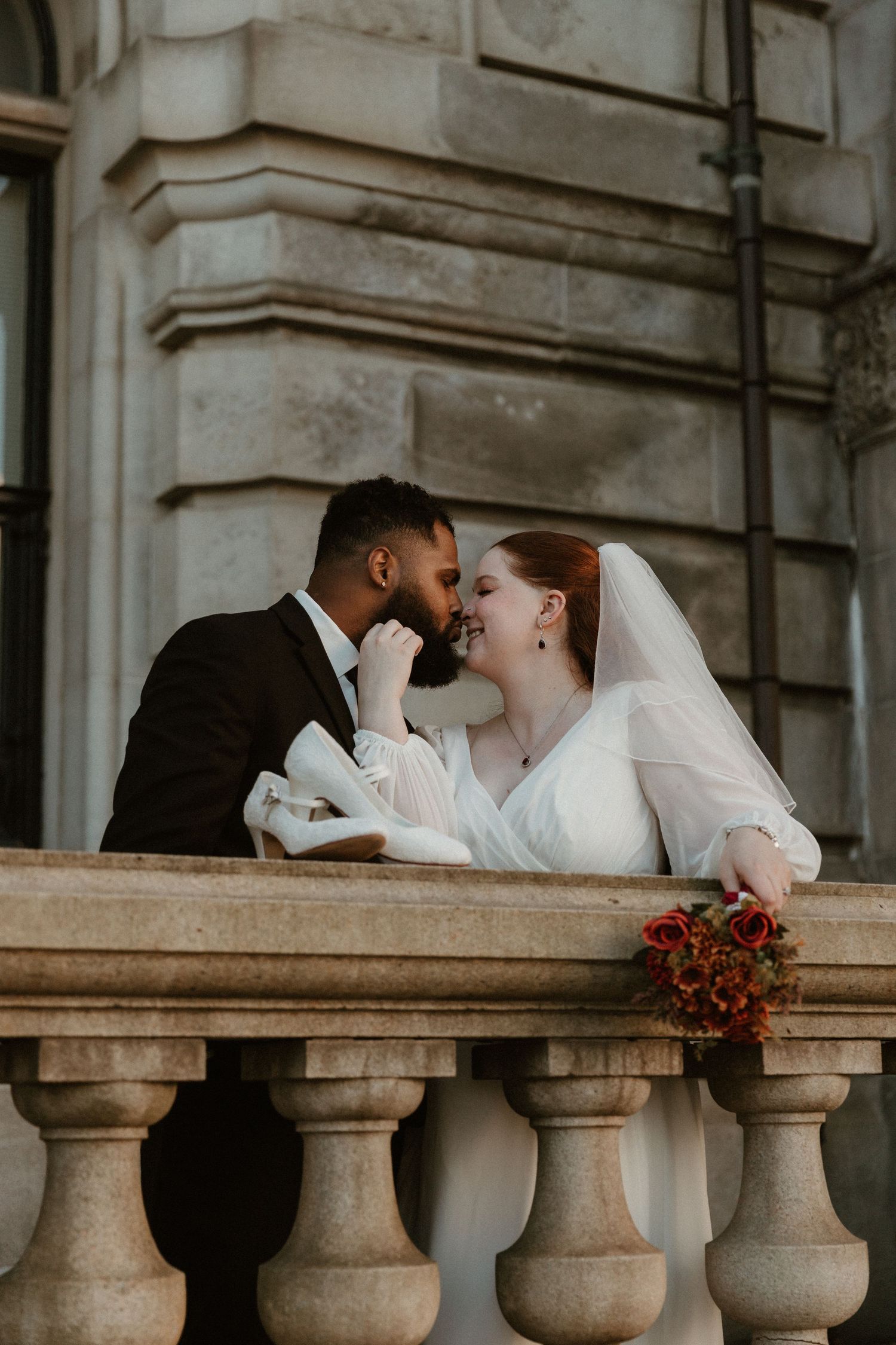 A romantic wedding couple shares an intimate moment on a stone balcony with red flowers.