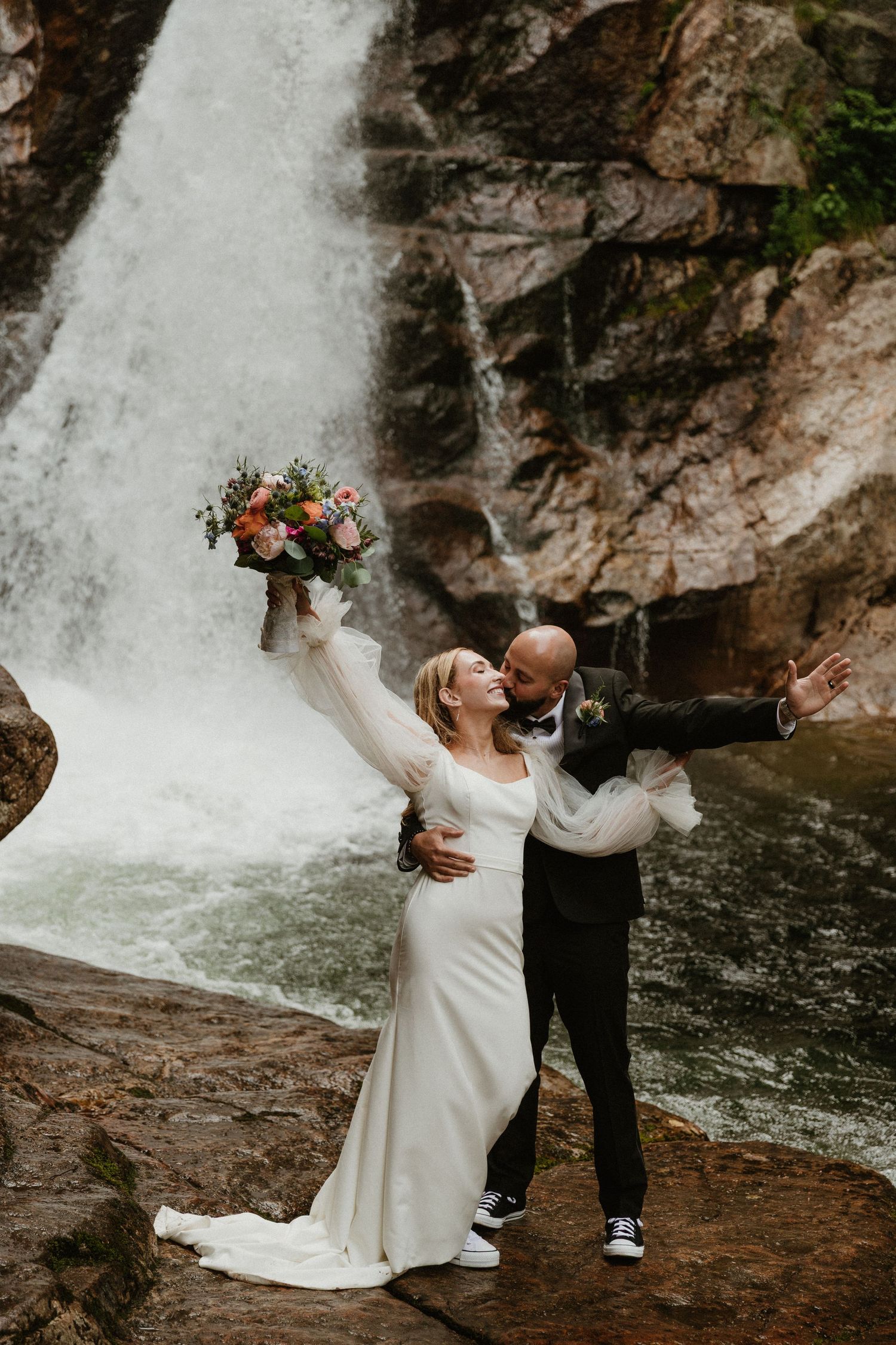 A romantic wedding portrait against a cascading waterfall backdrop with flowing white dress and floral bouquet.