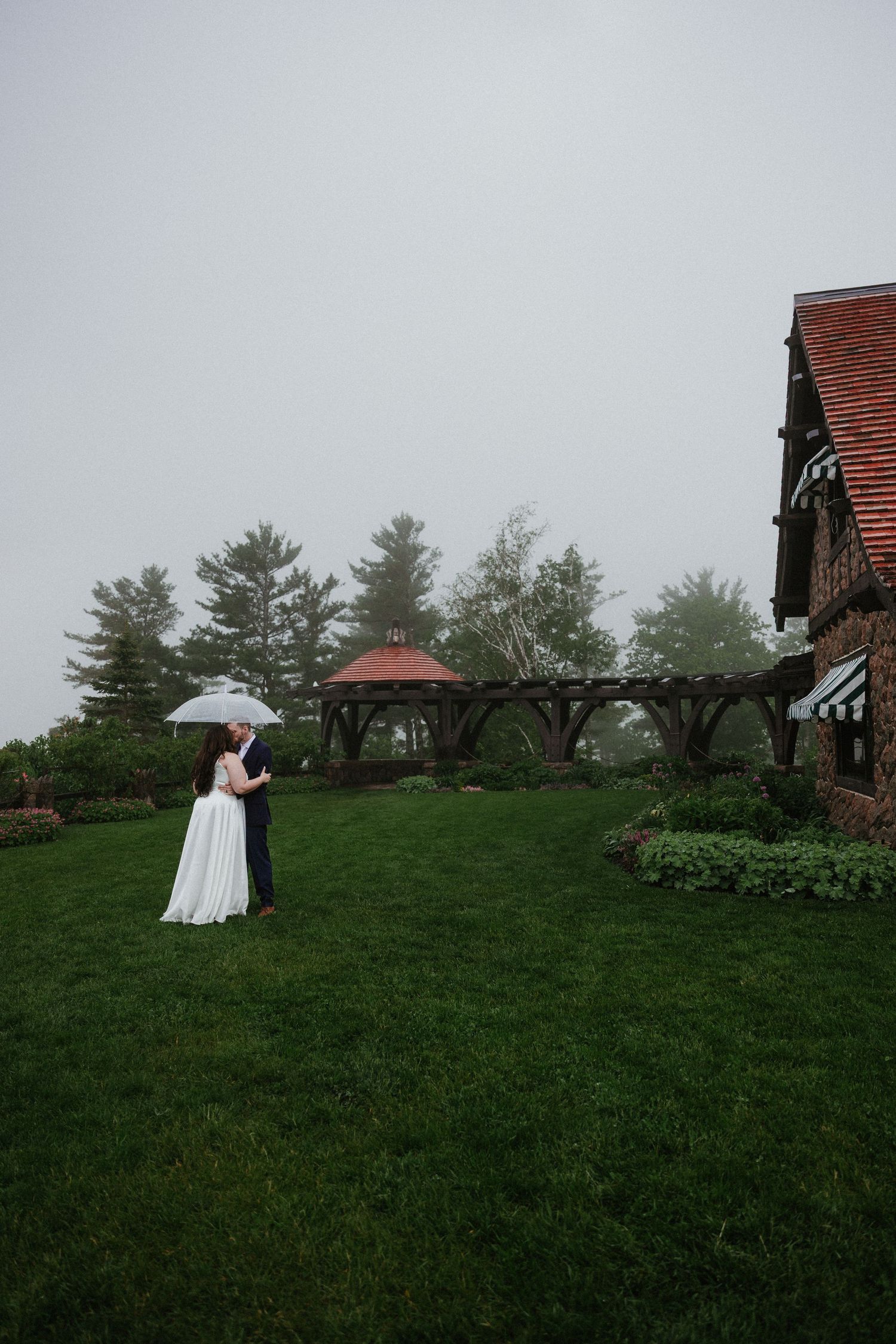 A couple shares a tender moment on a misty green hillside near a rustic building.